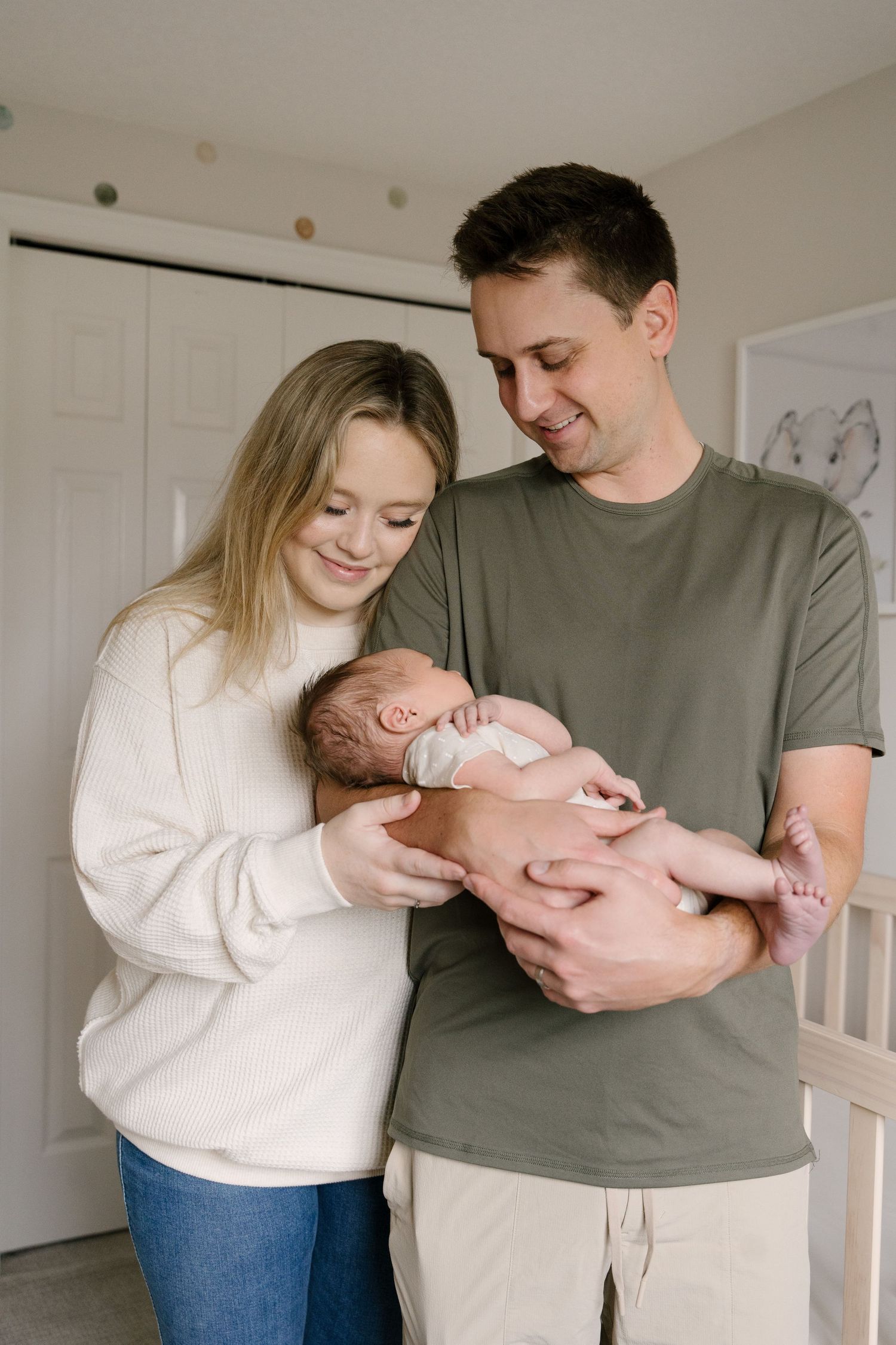 Happy new parents smiling and holding their newborn baby in a bright nursery room.