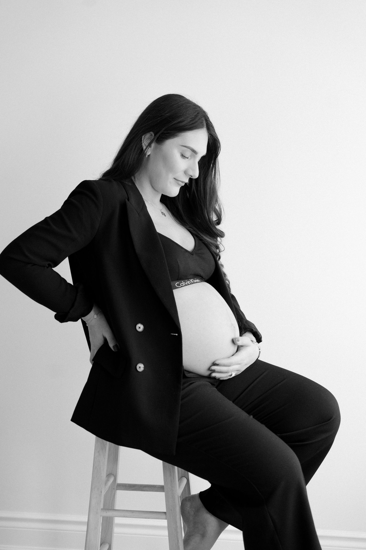 Elegant black and white maternity portrait of pregnant woman in blazer sitting on stool holding baby bump.