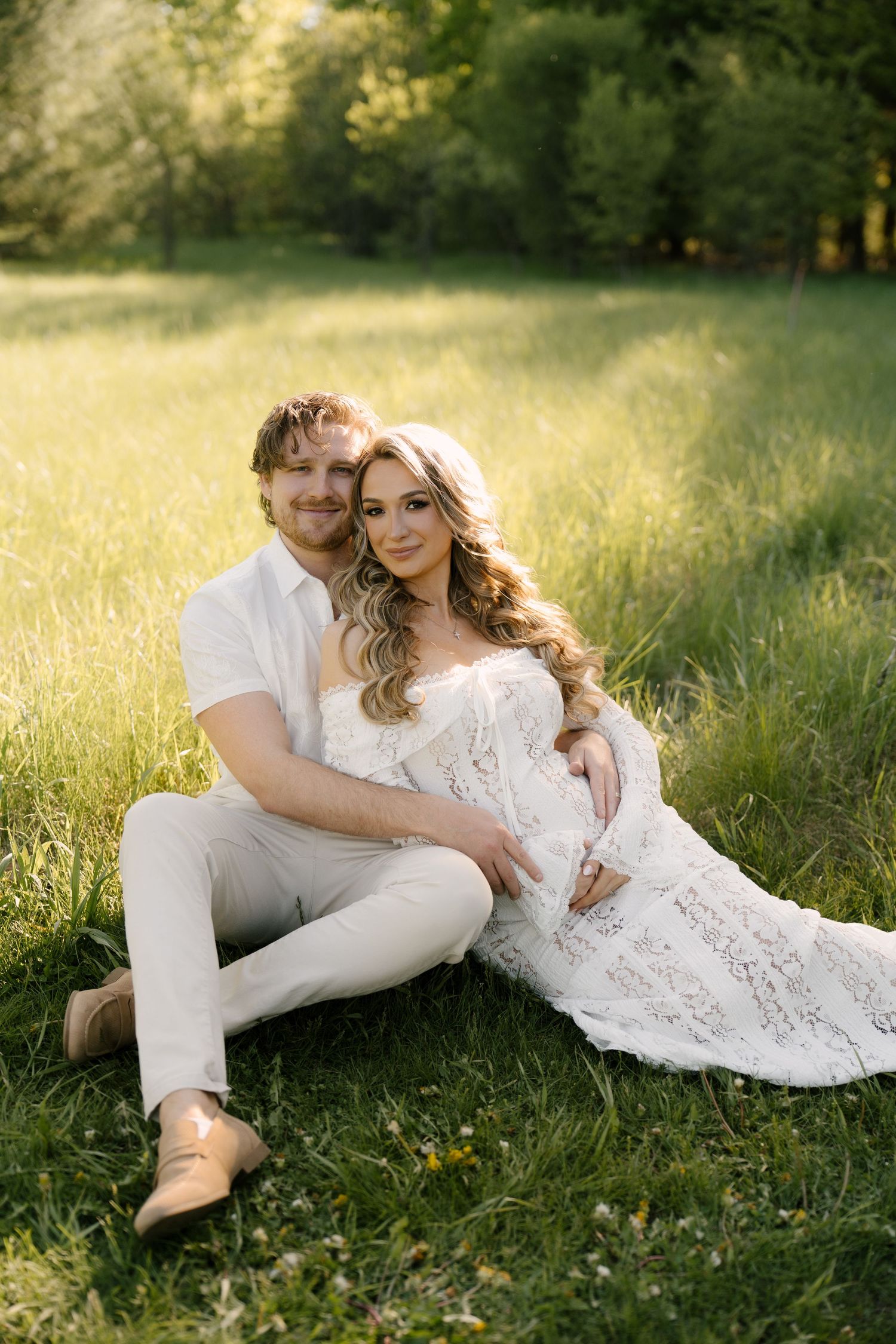 Couple sitting together on green grass in warm sunlight, woman in white lace maternity gown smiling.