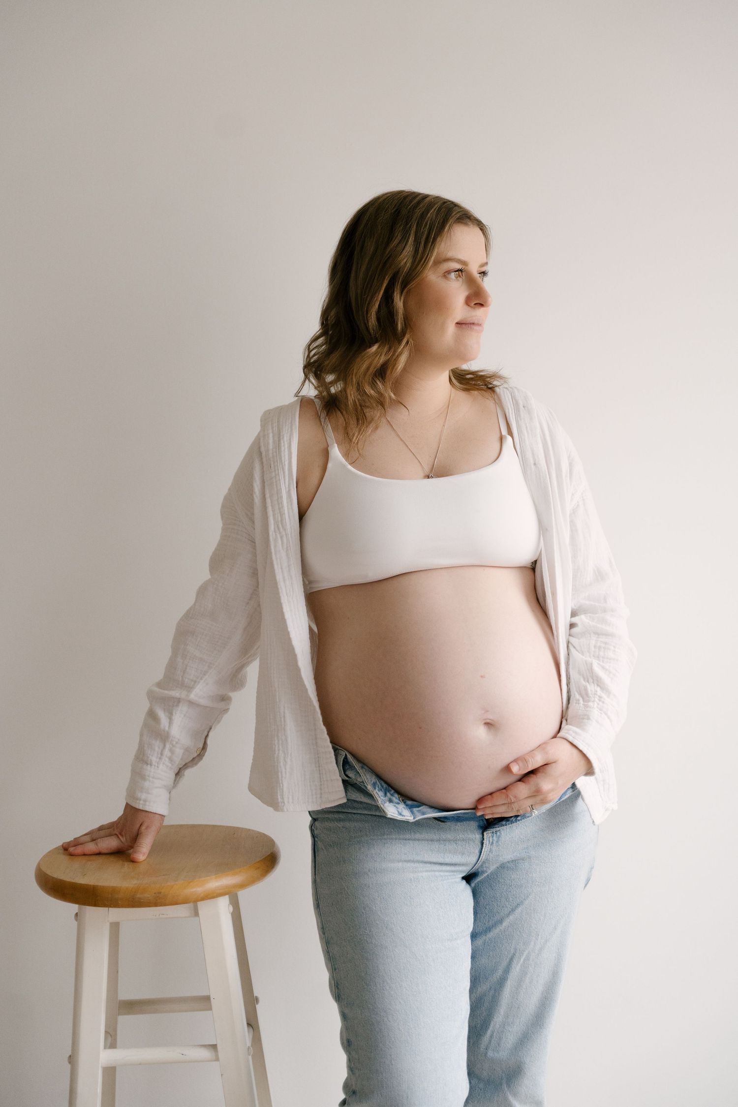 Pregnant woman in white bralette and open shirt standing beside a wooden stool, holding her belly.