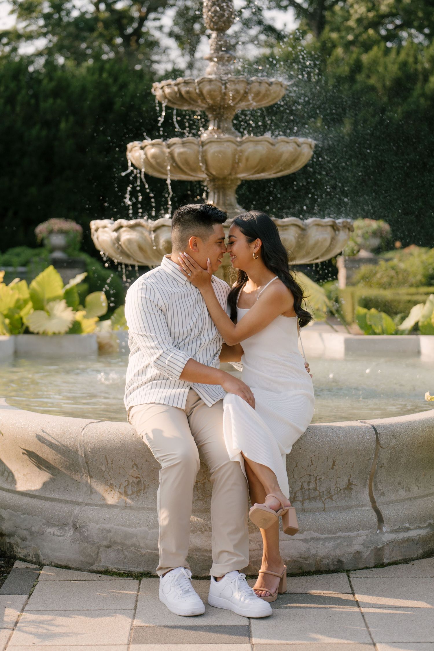 Couple sitting closely together on a stone fountain edge, almost kissing in a sunlit garden setting.