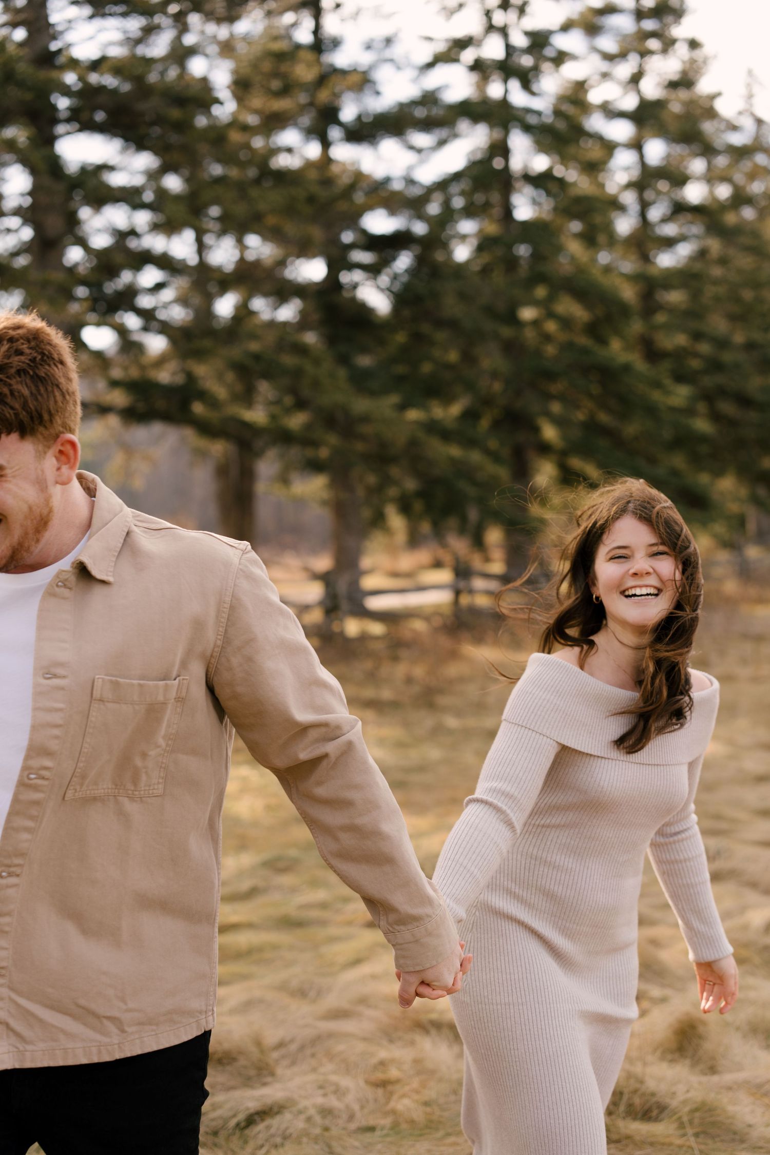 Laughing couple holding hands while walking outdoors in a pine forest, woman in a beige knit dress.