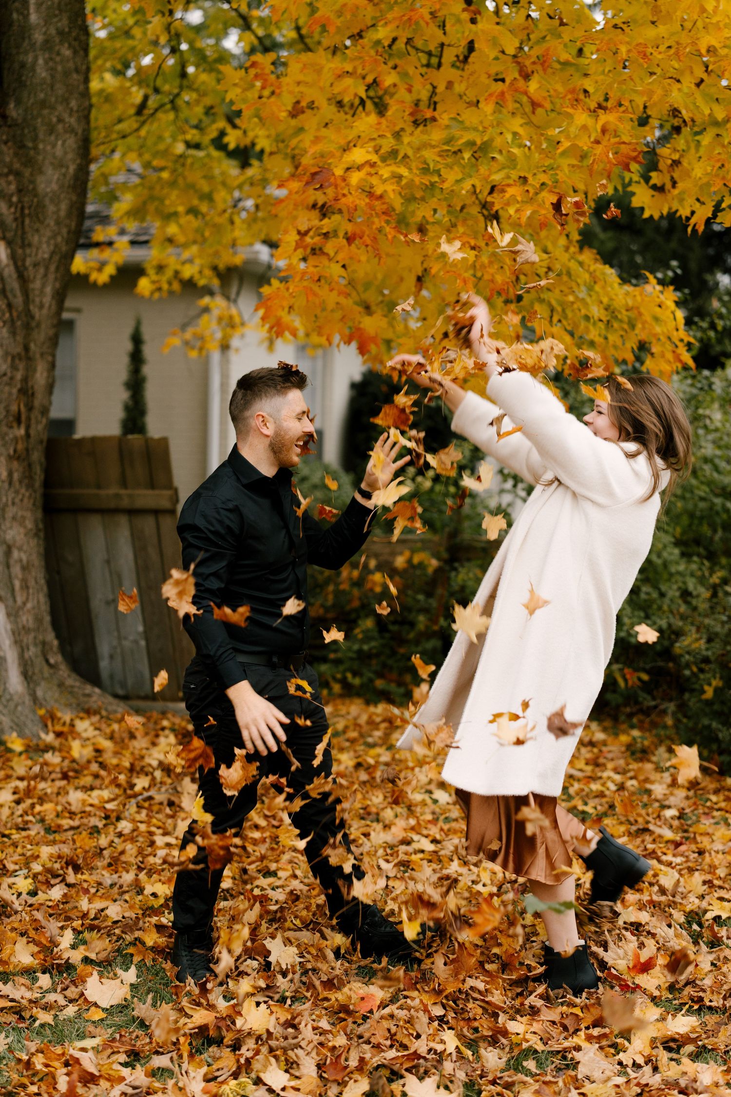 Laughing couple playfully throwing fall leaves at each other beneath a golden maple tree in autumn.