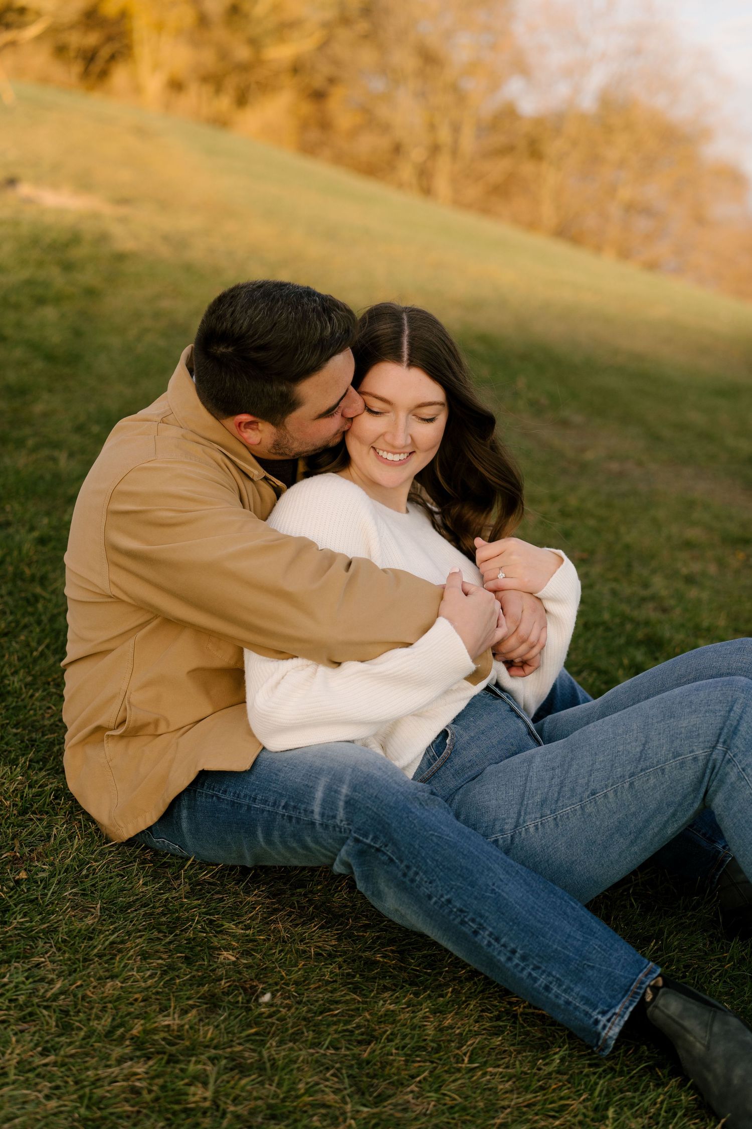 Romantic couple sitting on grass at sunset, man embracing woman from behind as she smiles joyfully.