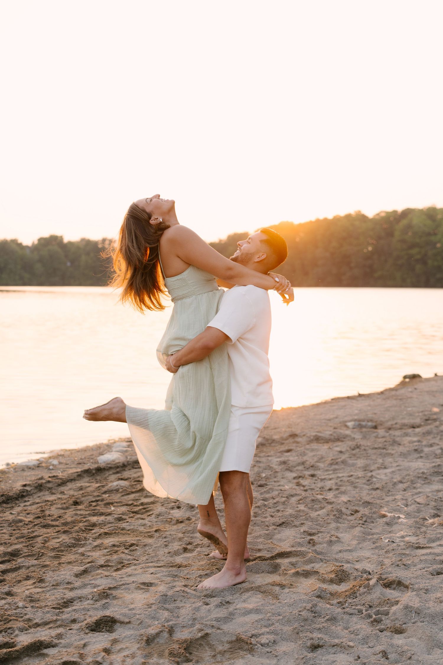 A man lifts and spins a laughing woman in a sage dress on a sandy beach at warm golden sunset.