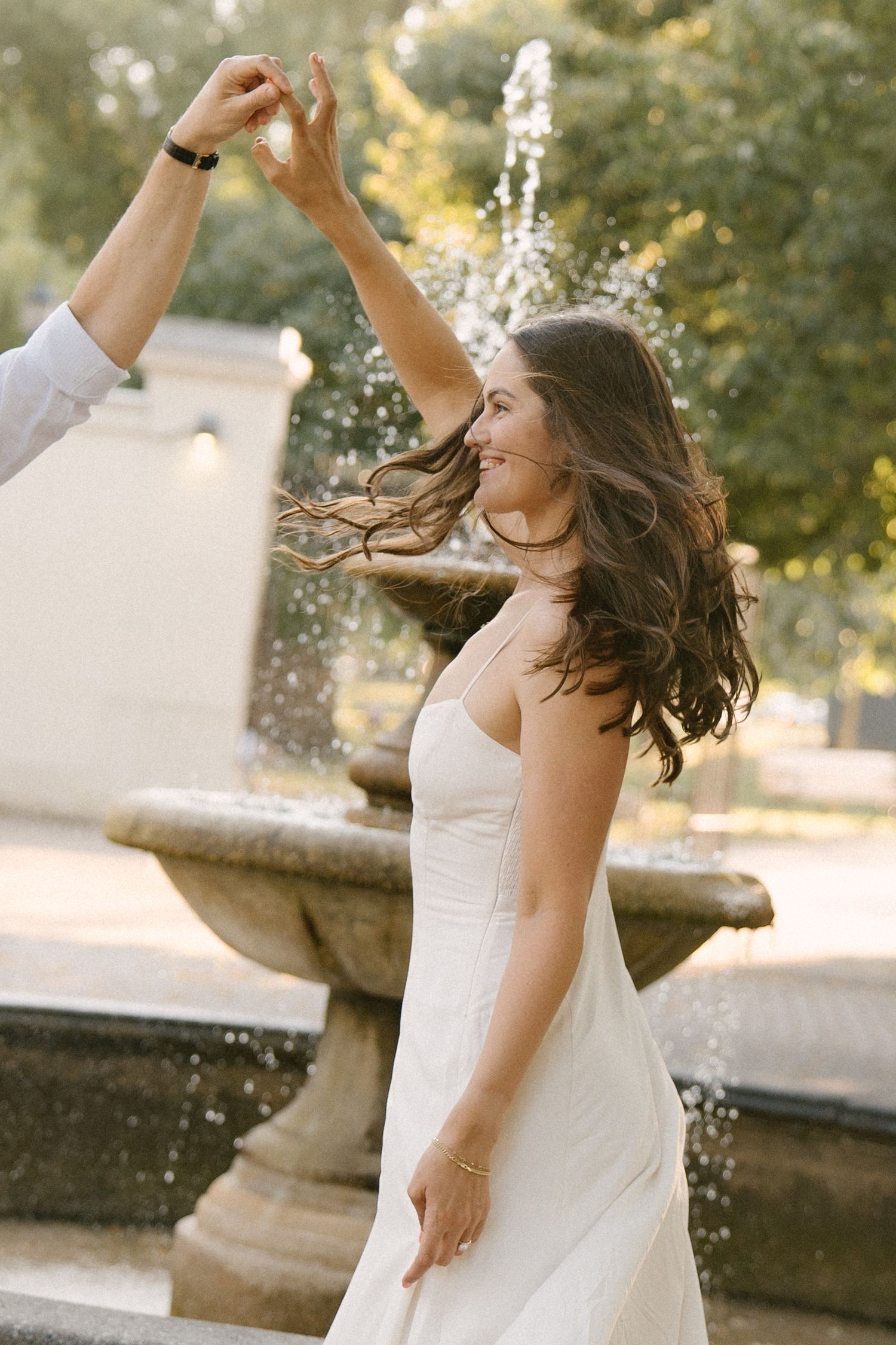 Woman in white dress spinning joyfully near a stone fountain in a sunny park.