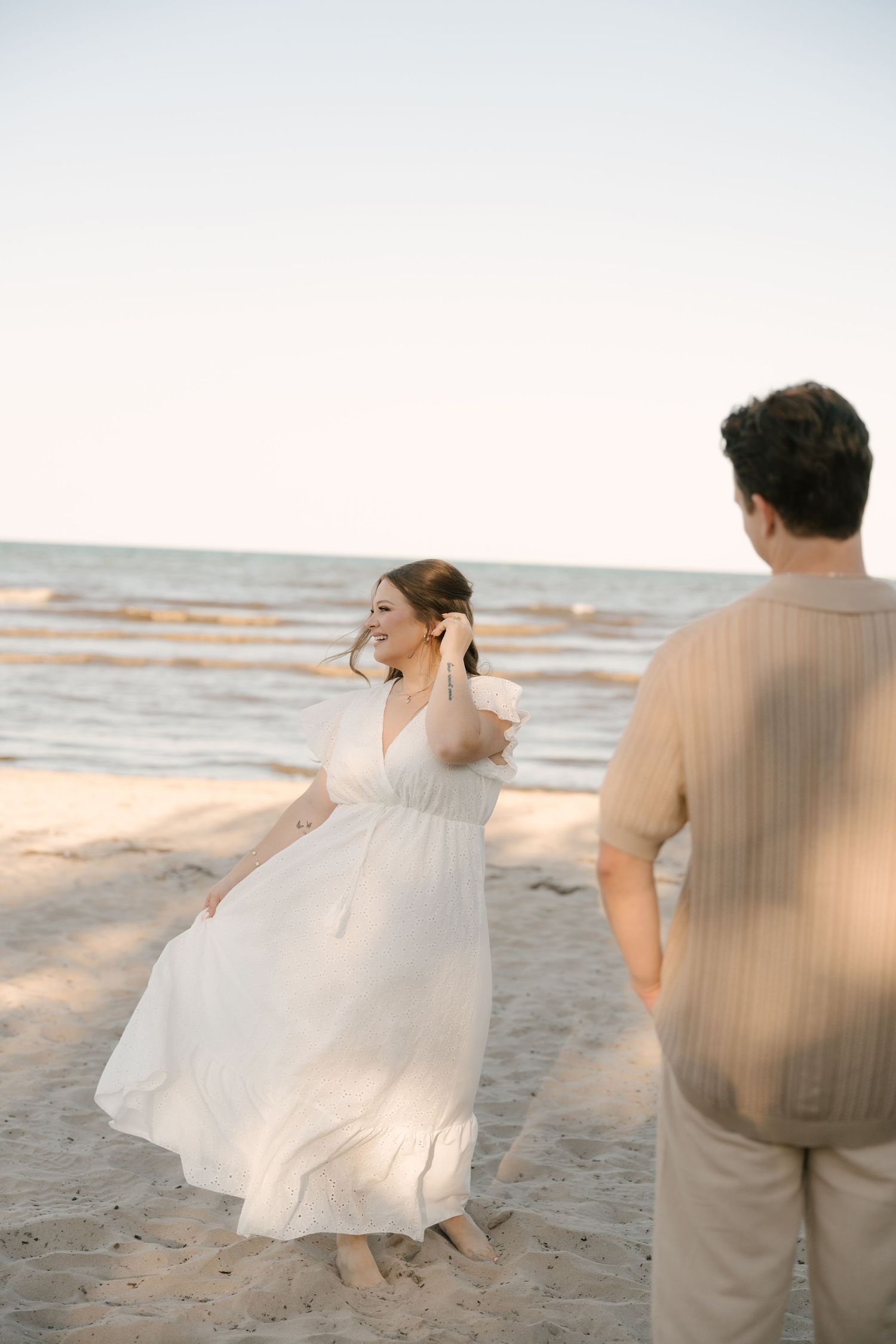 Smiling woman in white dress twirling on sandy beach at sunset with ocean backdrop.