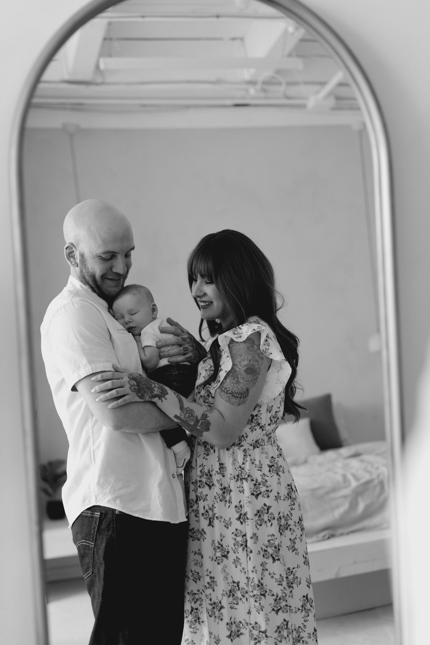 Black and white photo of tattooed parents holding newborn baby reflected in an arched mirror.