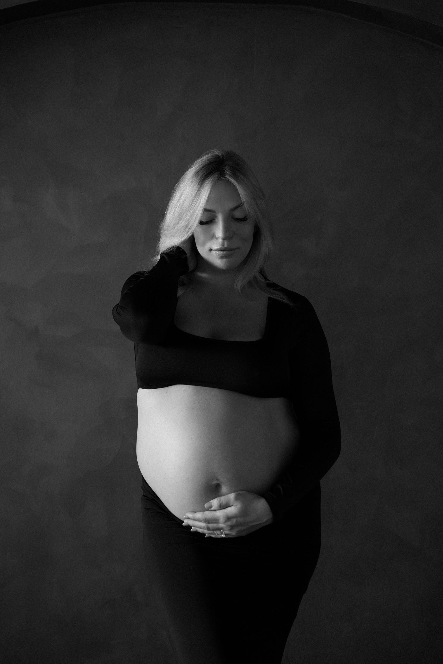 Black and white maternity photo of a blonde woman in a crop top cradling her baby bump against a dark background.