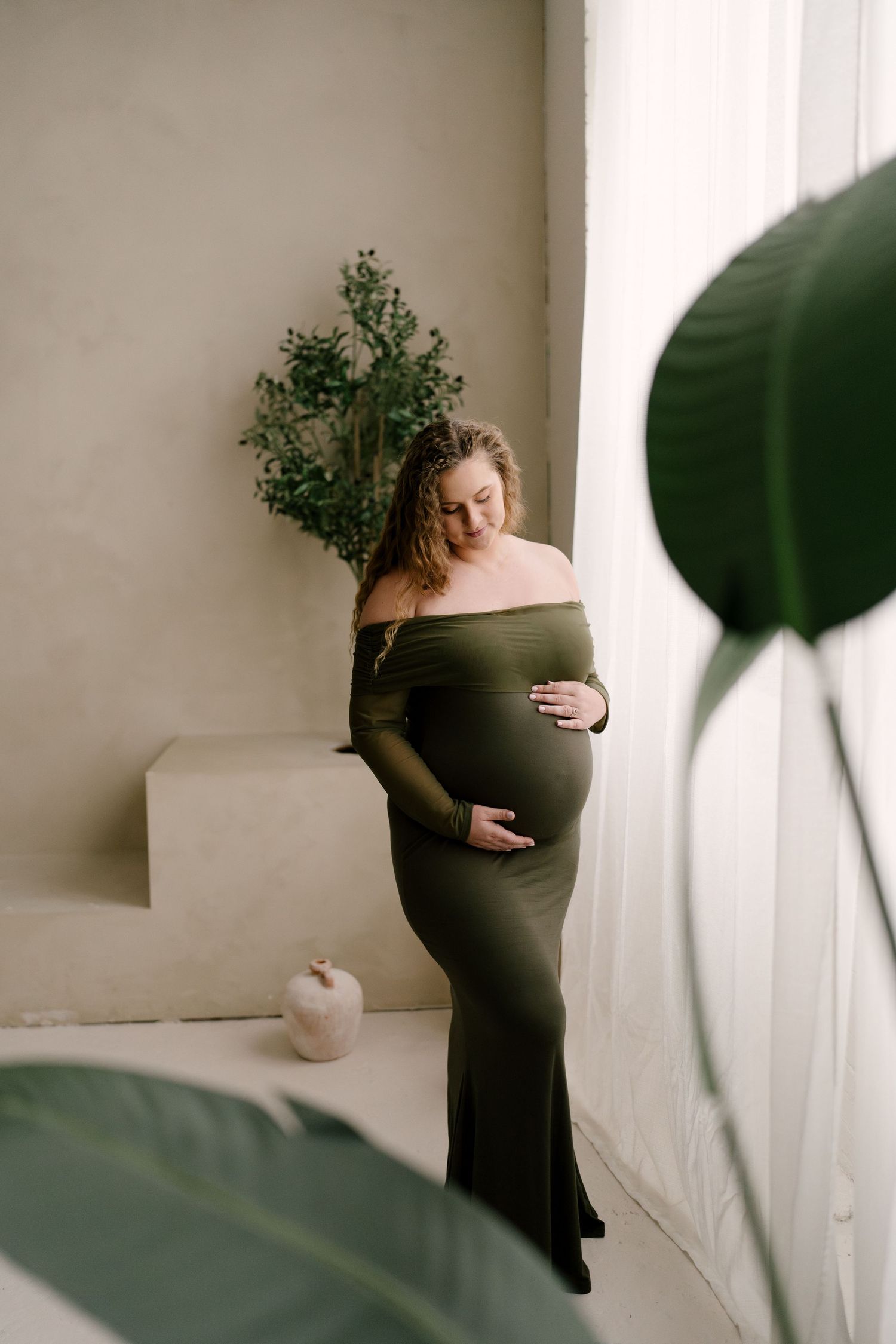 Pregnant woman in olive green off-shoulder maternity gown cradling baby bump in bright studio.