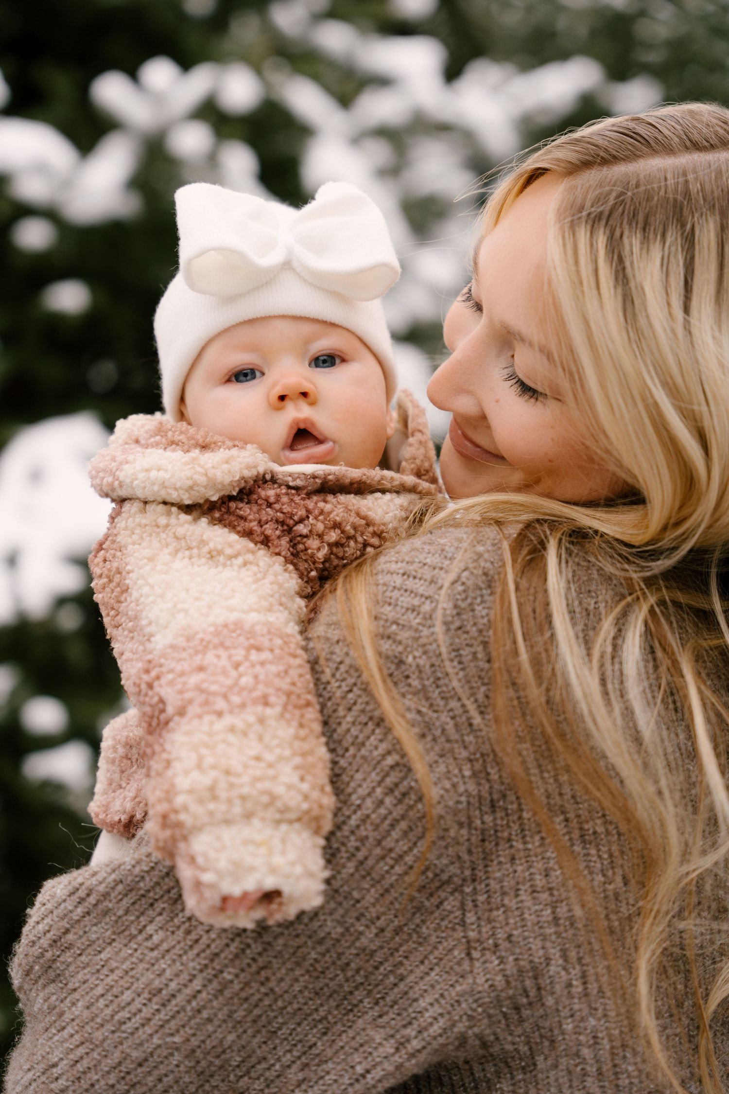 Mother holding blue-eyed baby girl in white bow hat and cozy winter outfit at snowy tree farm.