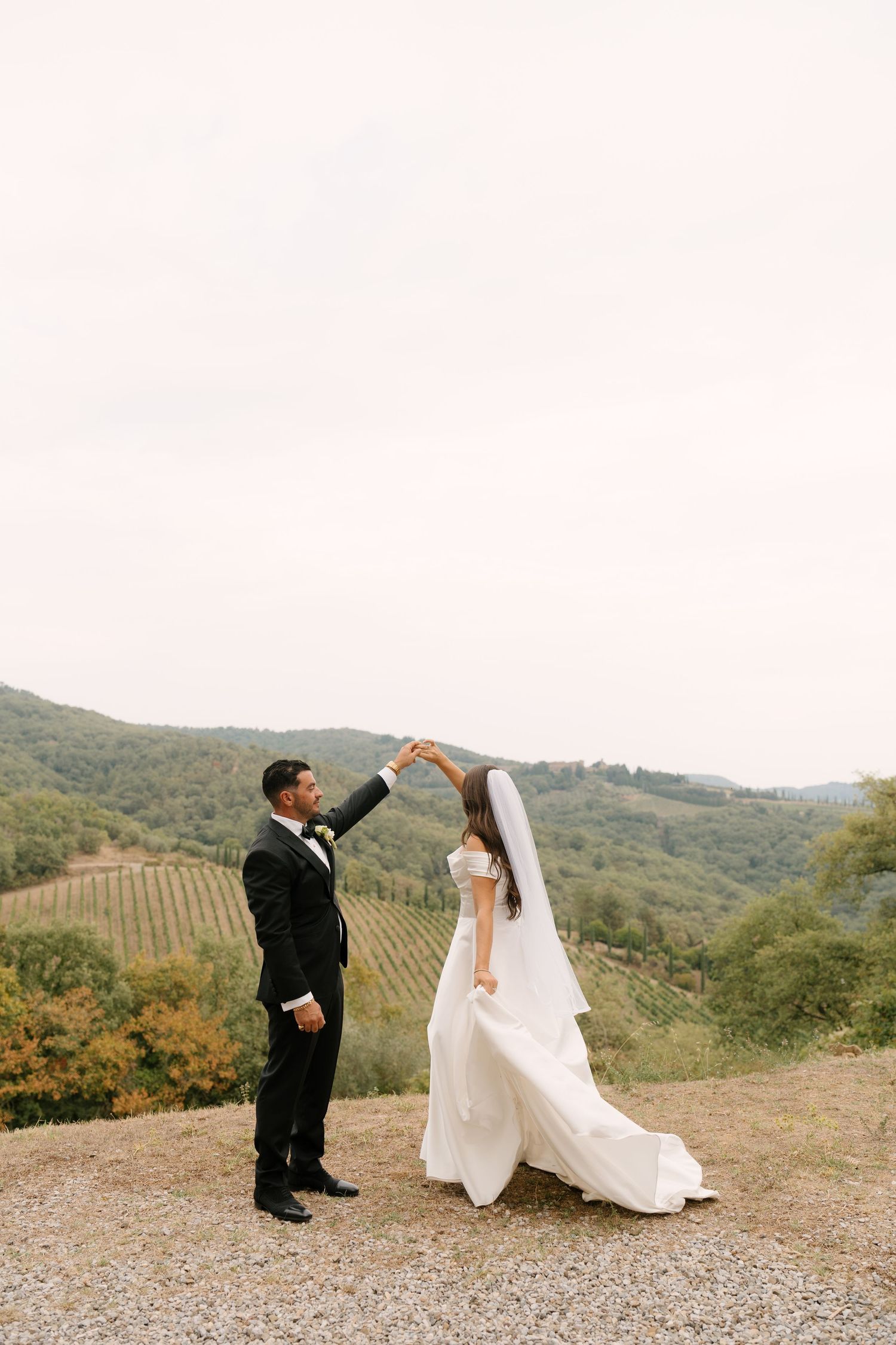 Groom twirls bride in white gown on hilltop overlooking stunning Tuscan vineyard landscape.