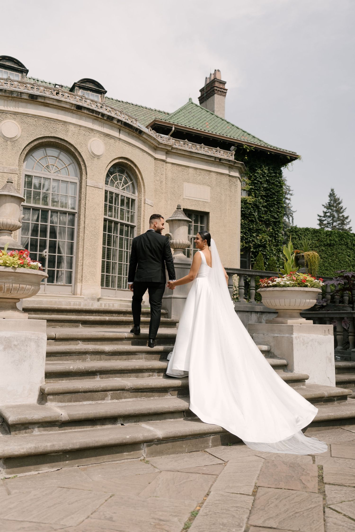 Bride and groom walking up stone steps of an elegant ivy-covered mansion, holding hands on their wedding day at Parkwood Estates.