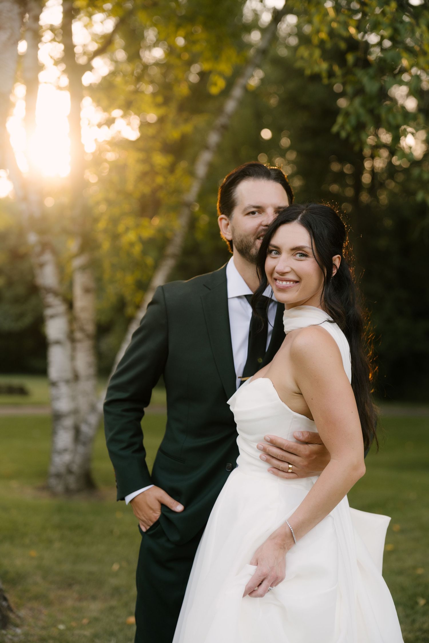 Bride and groom embracing at golden hour in a lush garden, groom in dark green suit, bride in white halter gown at Roseville Estates venue.
