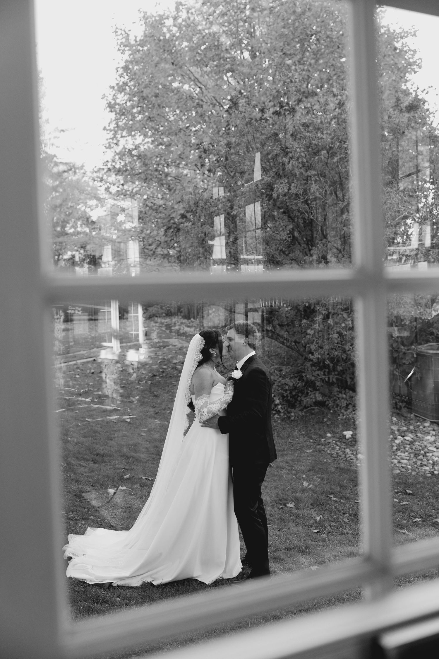 Black and white wedding photo of bride and groom kissing outdoors by a pond, captured through a window.