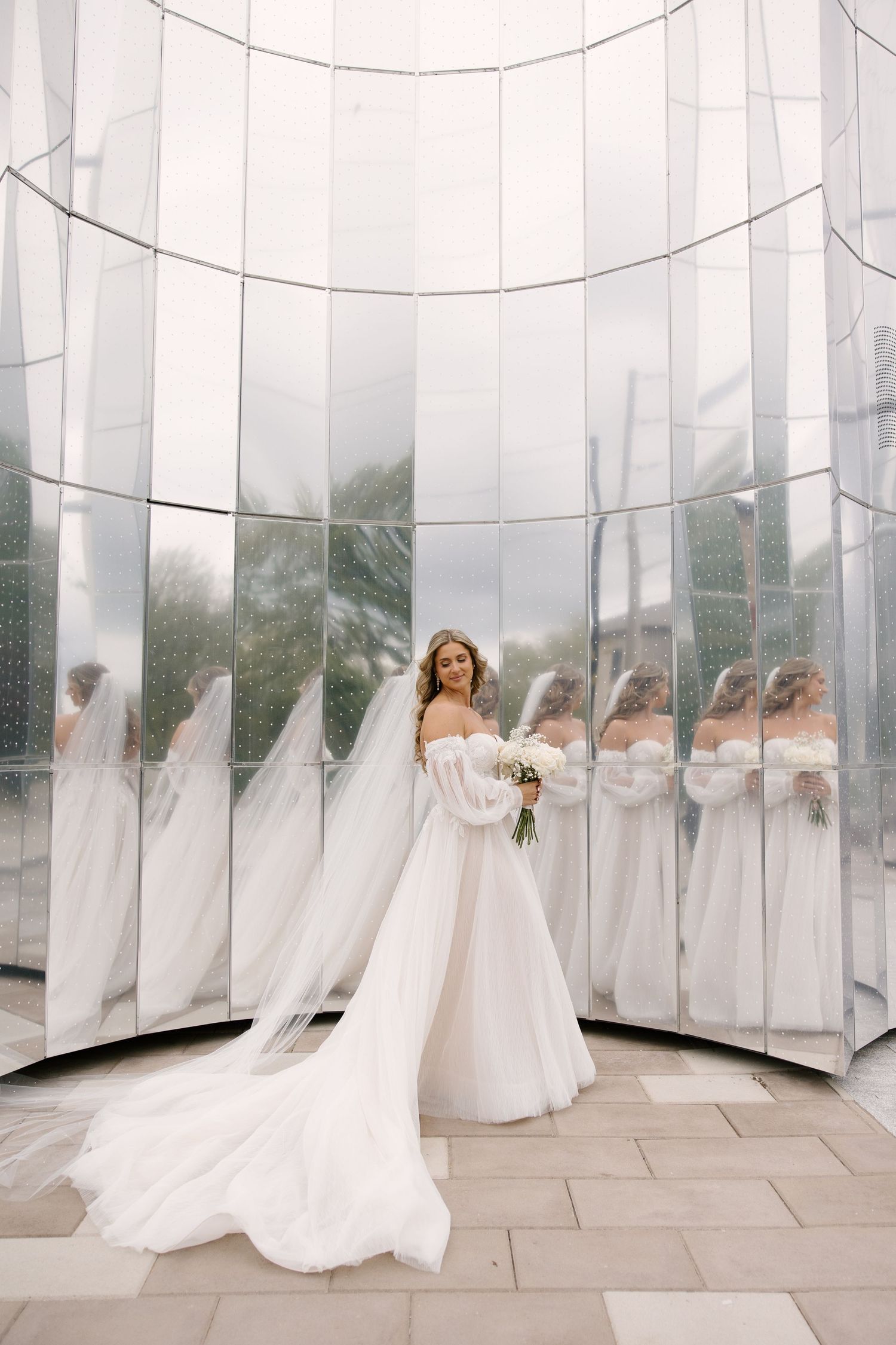 Bride in strapless white gown with long veil poses before mirrored glass wall with bouquet at Tapestry Hall wedding venue.