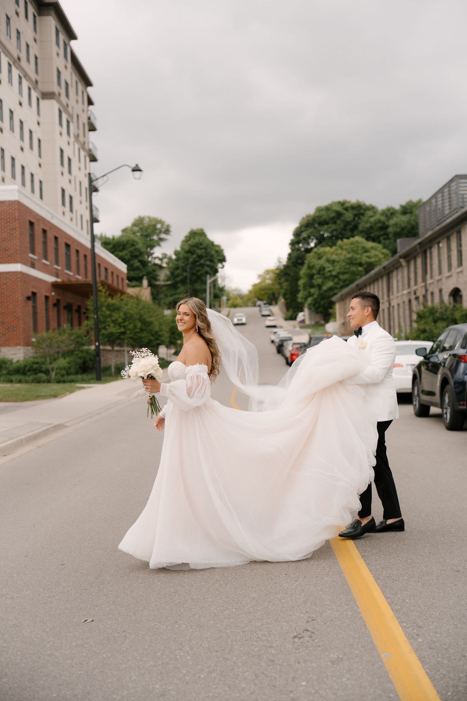 Groom lifts bride's flowing white veil as couple crosses a street in an urban setting at Tapestry Hall wedding venue.