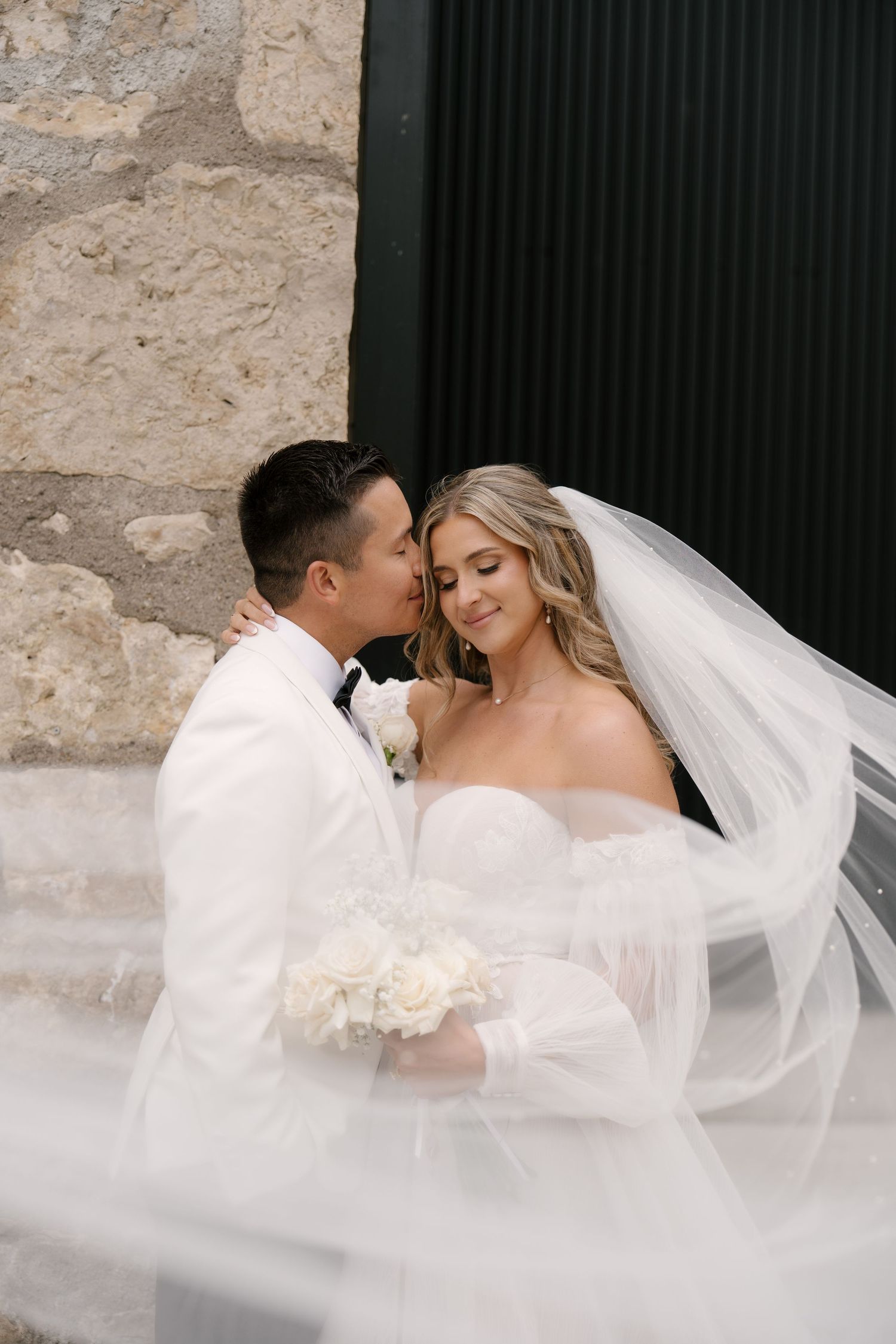 Groom kisses bride's cheek as her veil billows around them against a rustic stone backdrop outside Tapestry Hall.