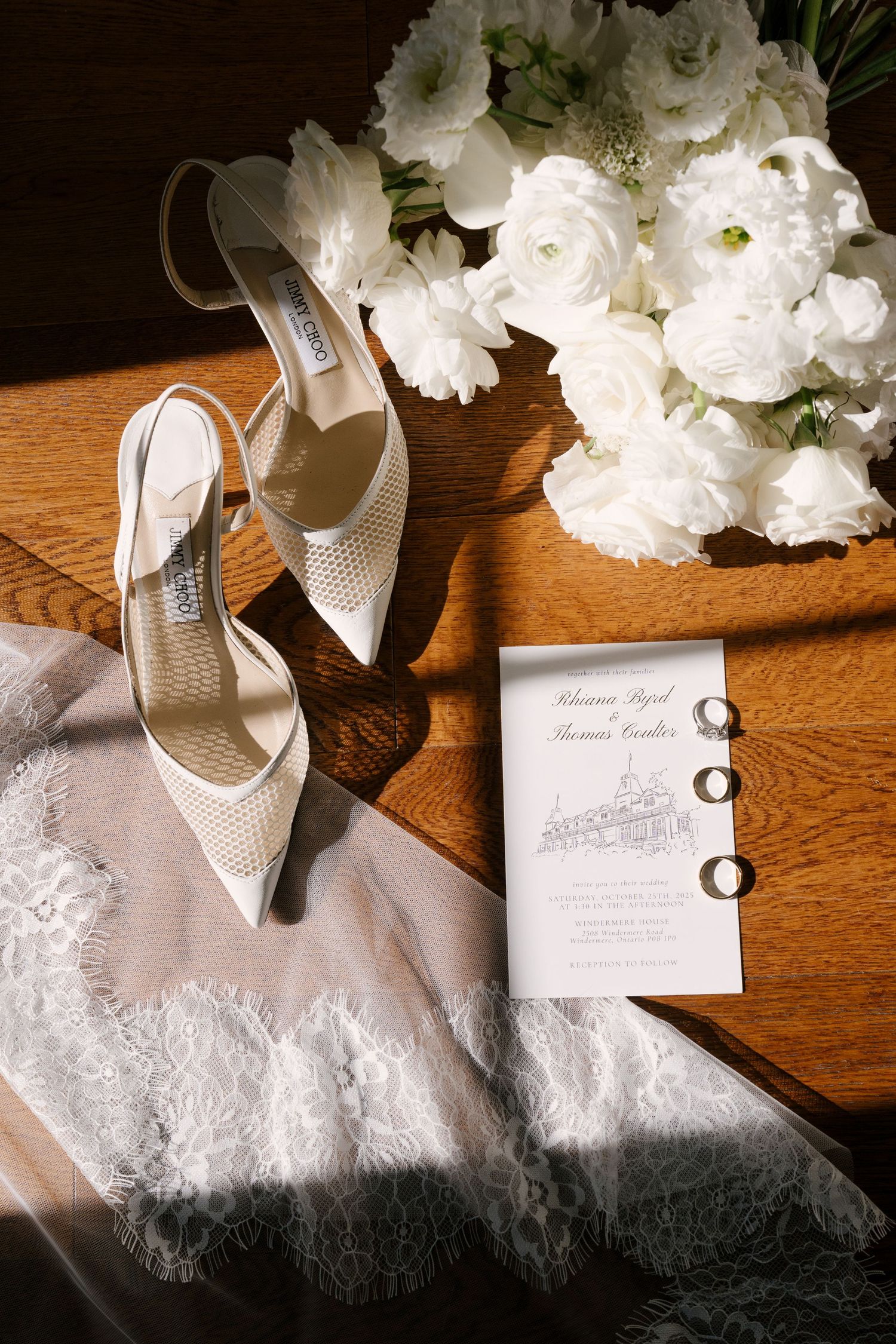 White Jimmy Choo wedding heels, lace veil, floral bouquet, and wedding invitation with rings on wooden table.