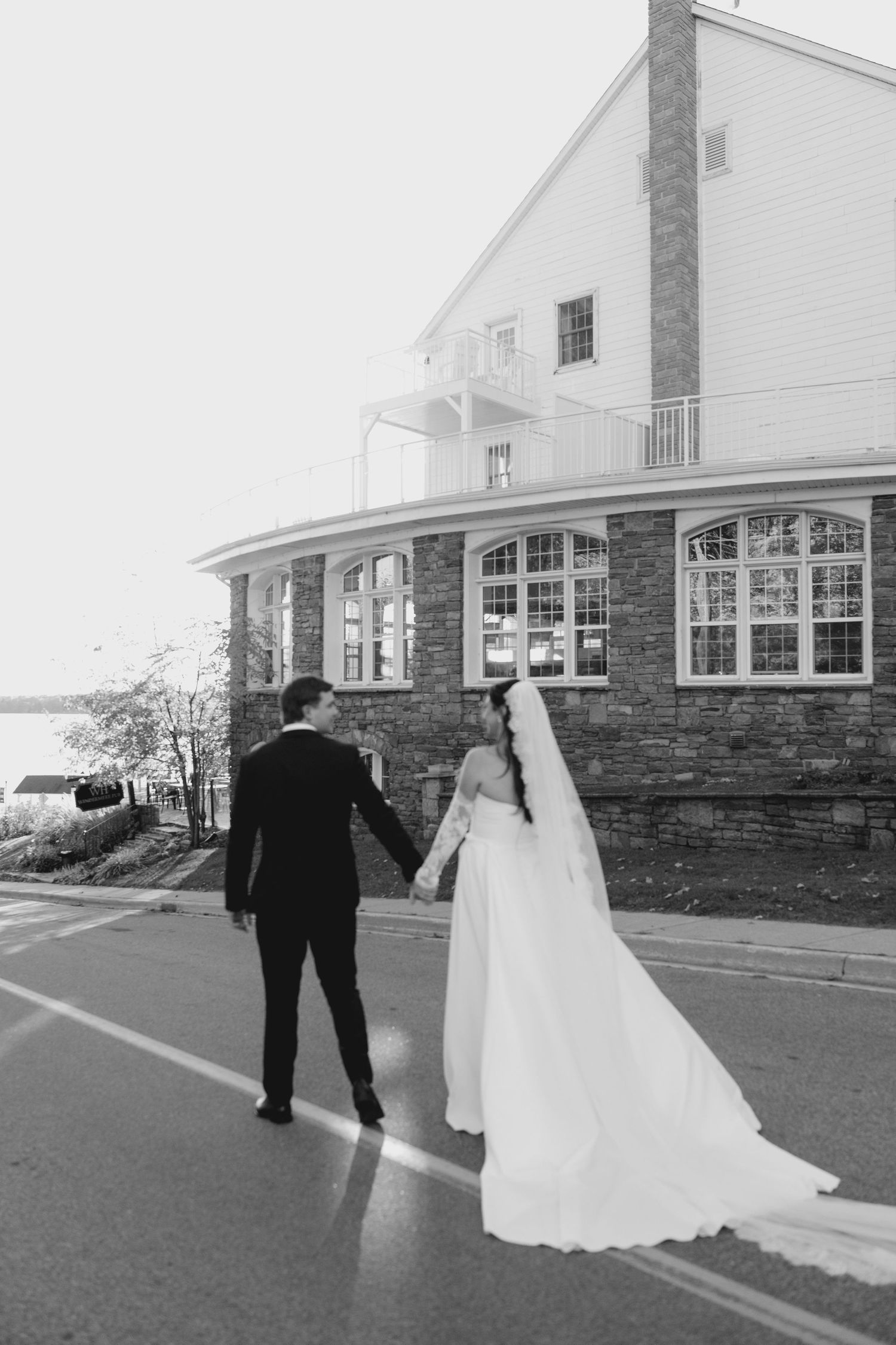 Black and white photo of bride and groom holding hands walking toward a lakeside wedding venue.