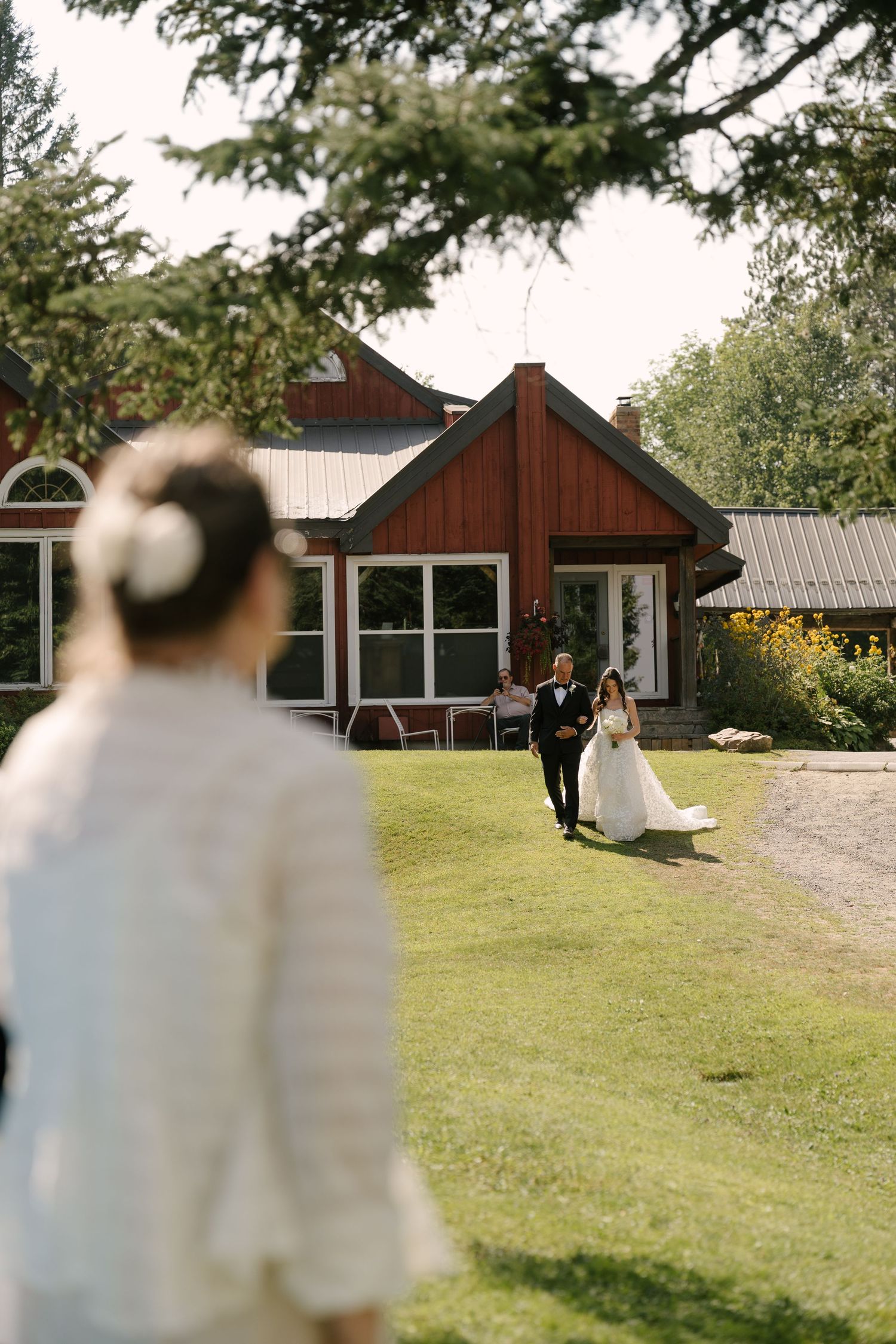 Bride and groom walk across green lawn toward wedding guest near red barn venue on sunny day.