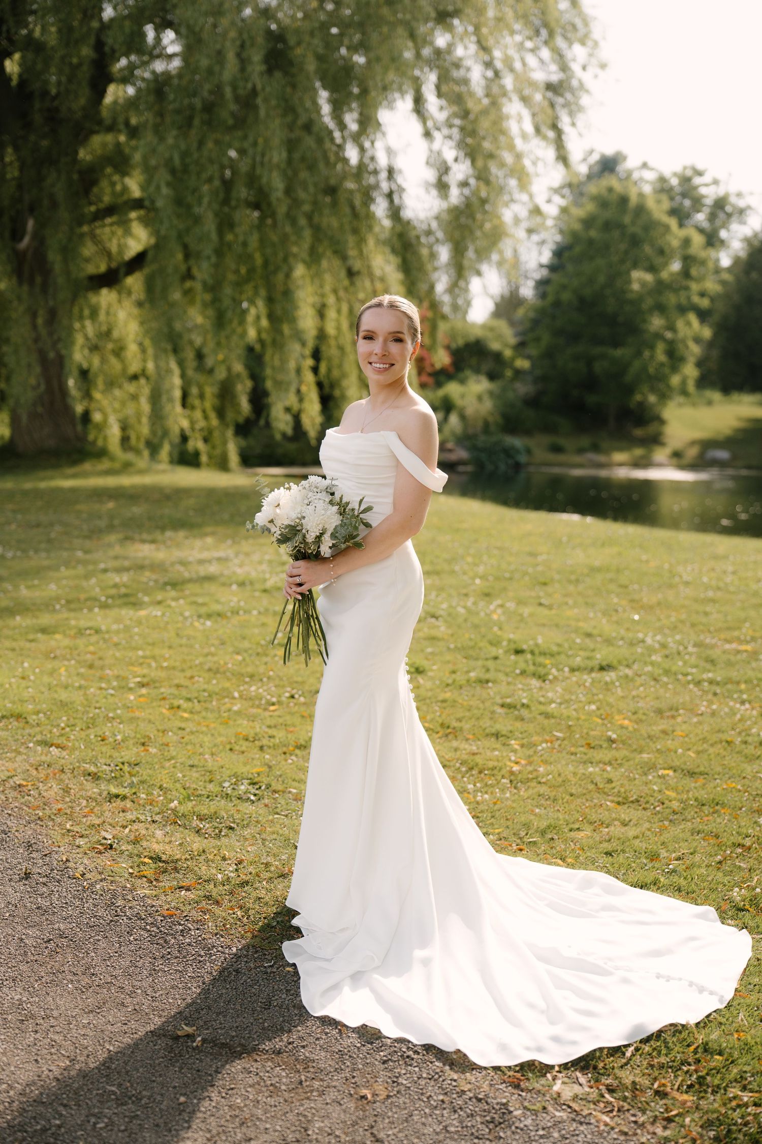 Elegant bride in off-shoulder mermaid gown holds white bouquet in lush green park setting.