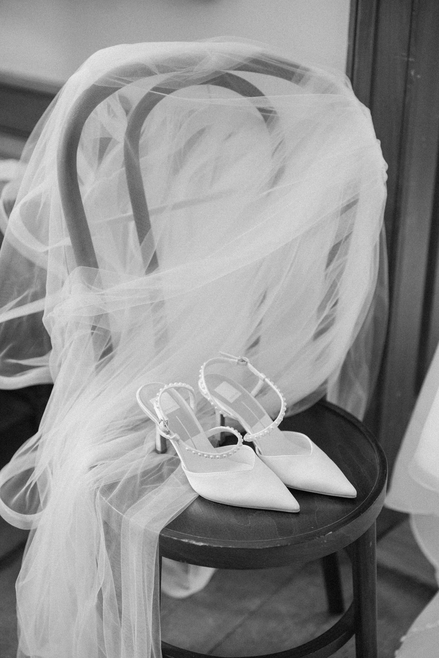 Black and white photo of white bridal heels and veil draped over a wooden chair.