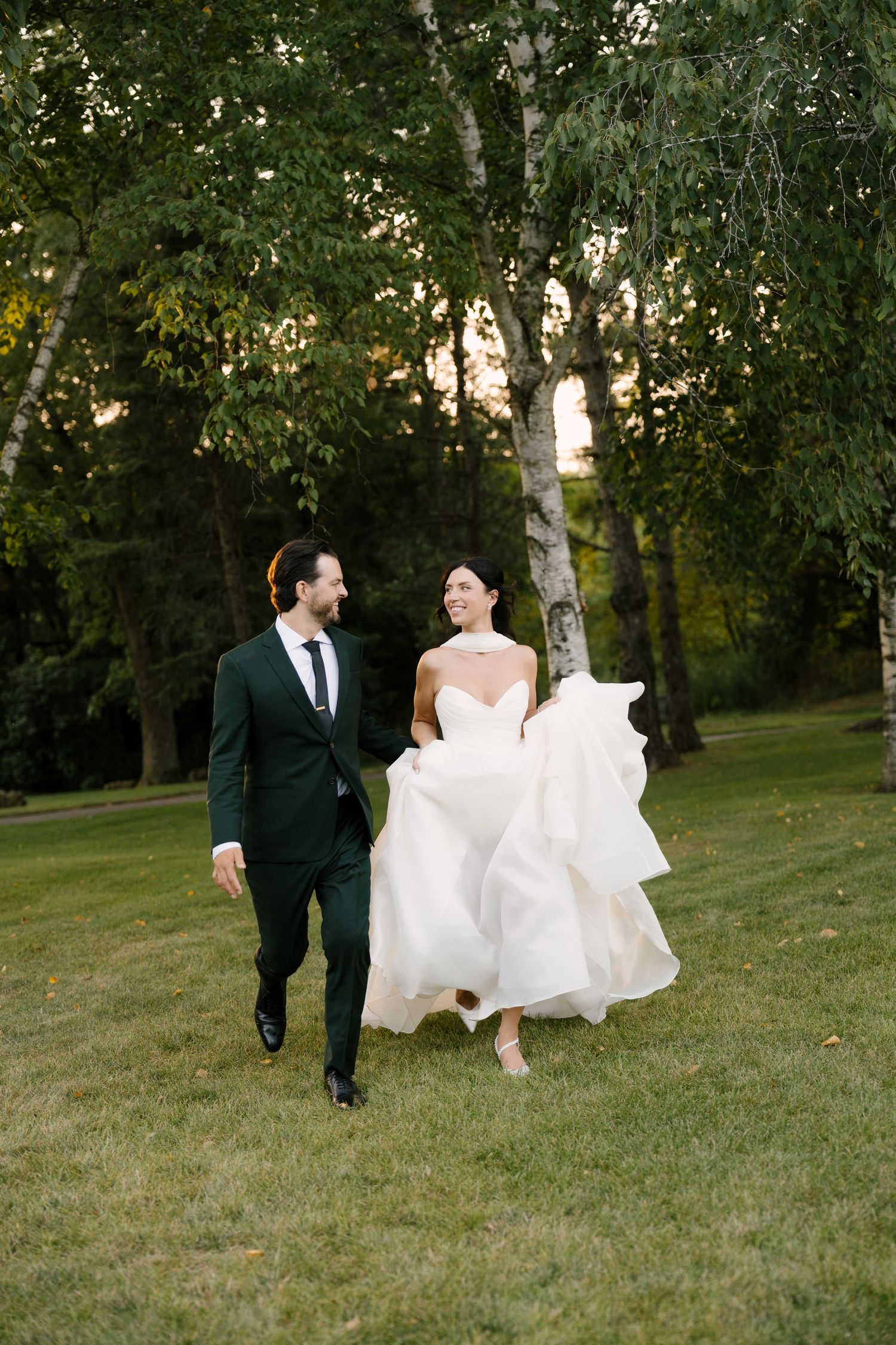 Bride and groom walking hand in hand through a sunlit garden, bride in strapless white gown, groom in green suit at Roseville Estates.