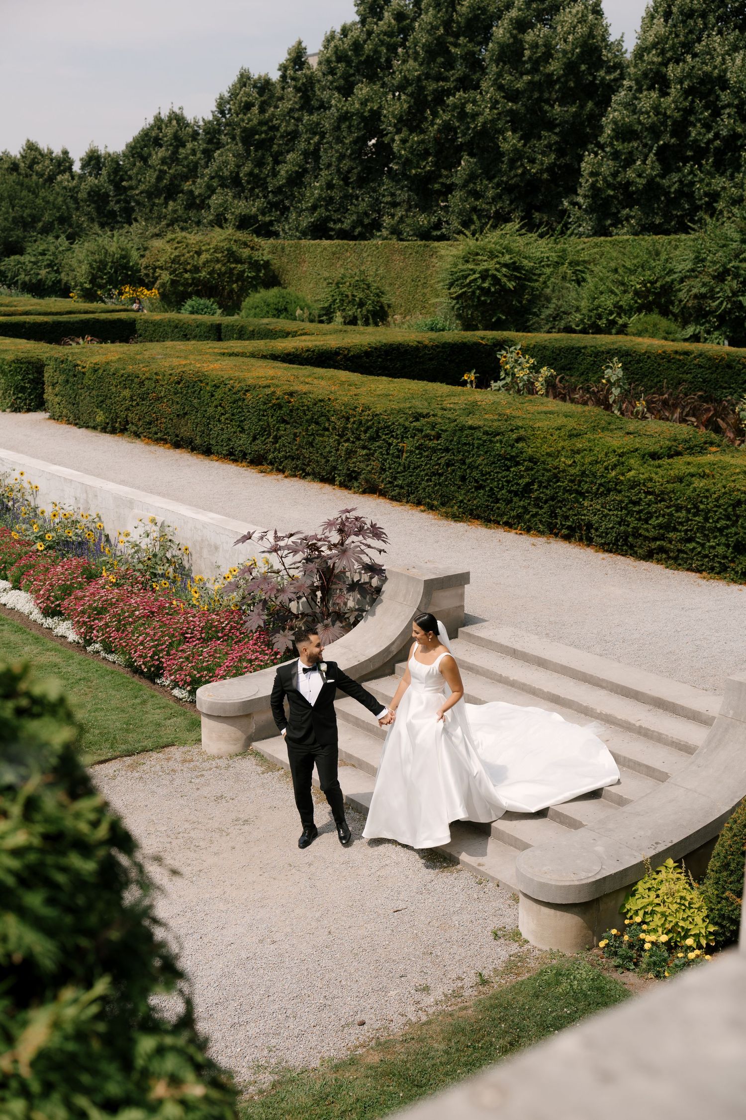 Aerial view of bride and groom holding hands on curved garden steps surrounded by manicured hedges and flowers at Parkwood Estates.