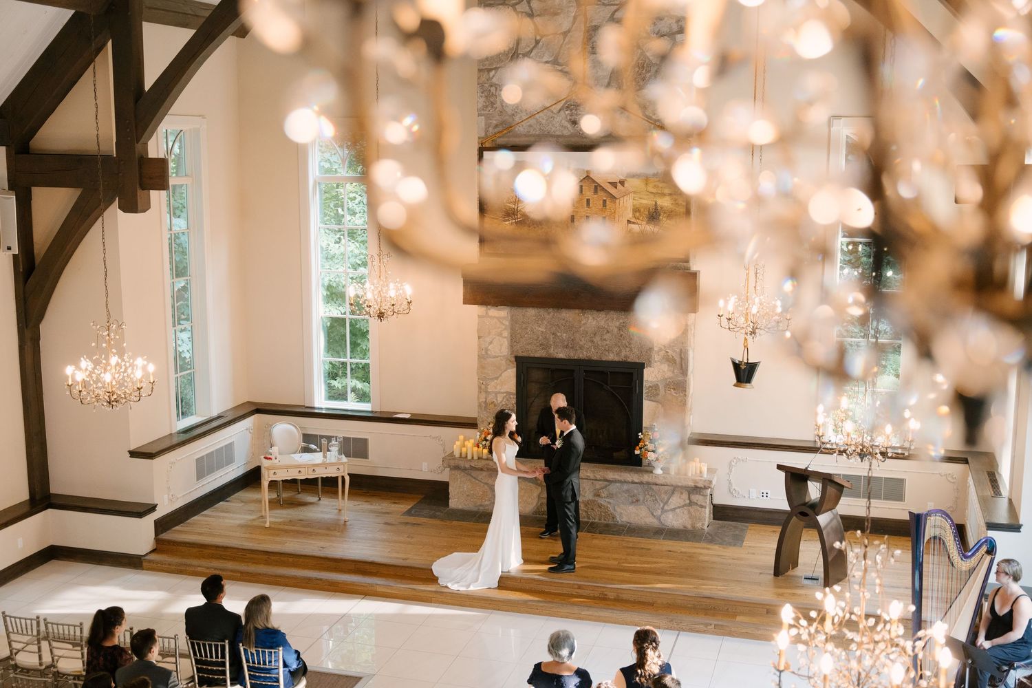 Elegant wedding ceremony in a bright Ancaster Mill chapel with chandeliers, stone fireplace, and couple exchanging vows on a wooden floor.