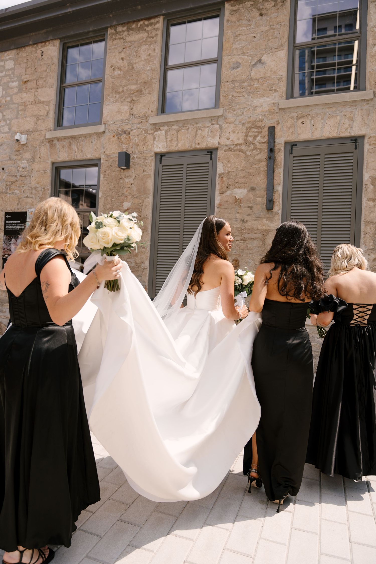 Bridesmaids in black gowns help carry the bride's white wedding dress train outside a stone Tapestry Hall building.
