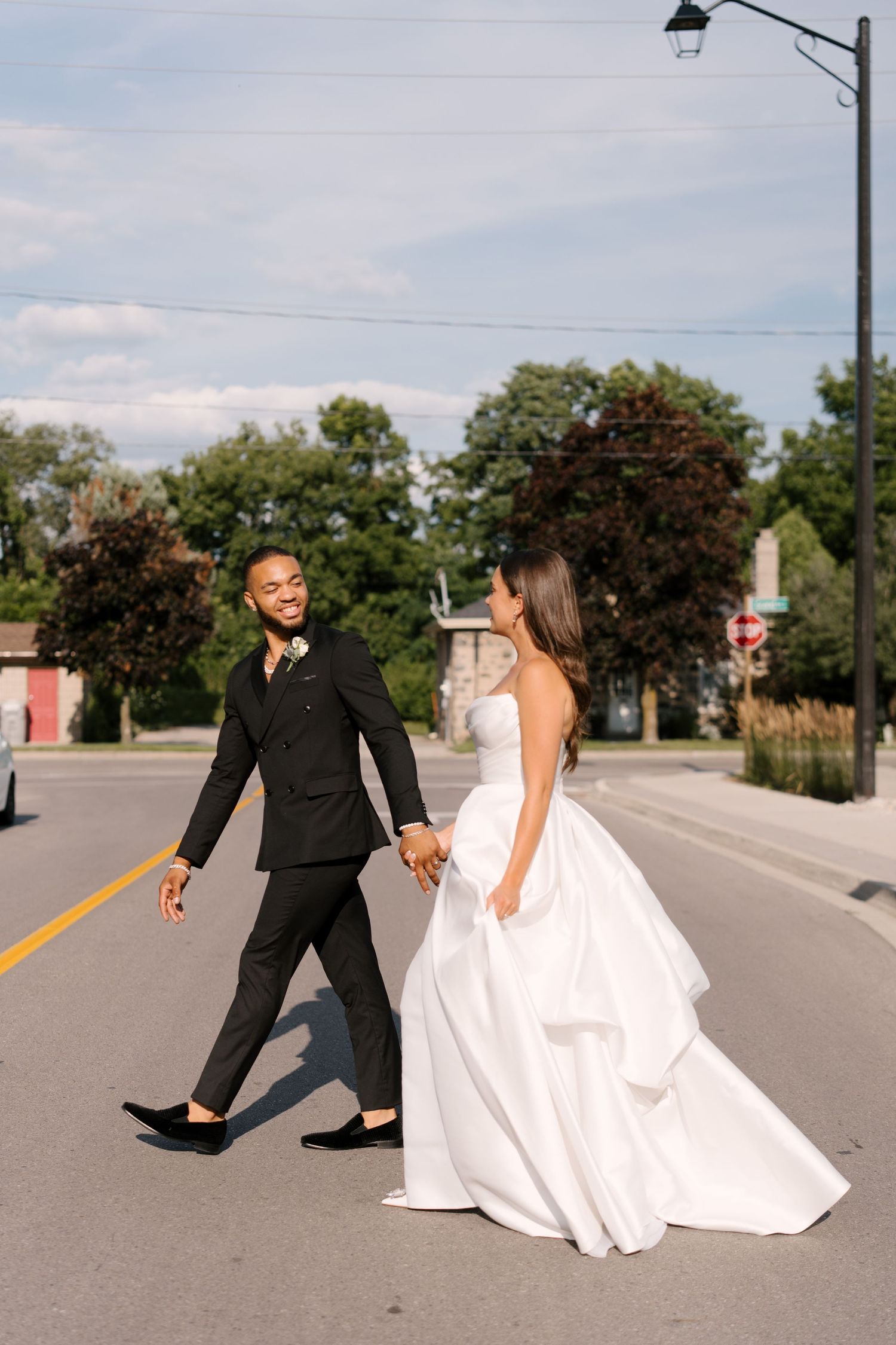 Bride in strapless white ballgown and groom in black suit hold hands crossing a sunny street.