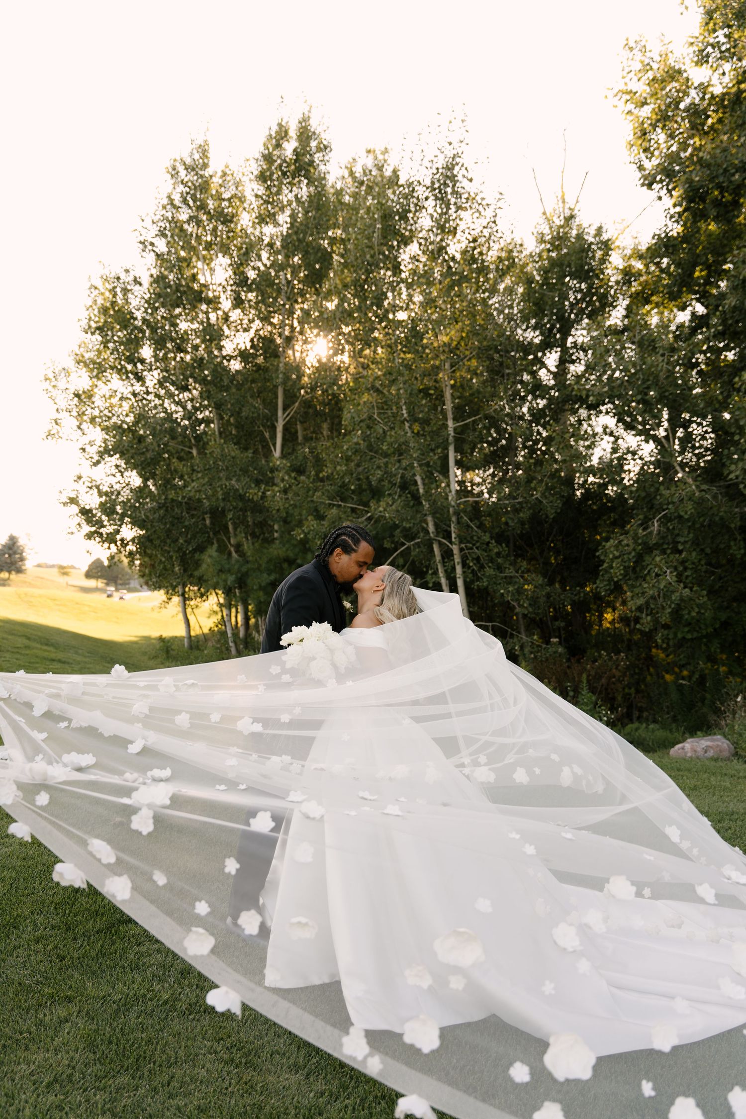Bride and groom kiss outdoors at sunset with floral veil dramatically spread across lawn at Whistle Bear wedding venue.