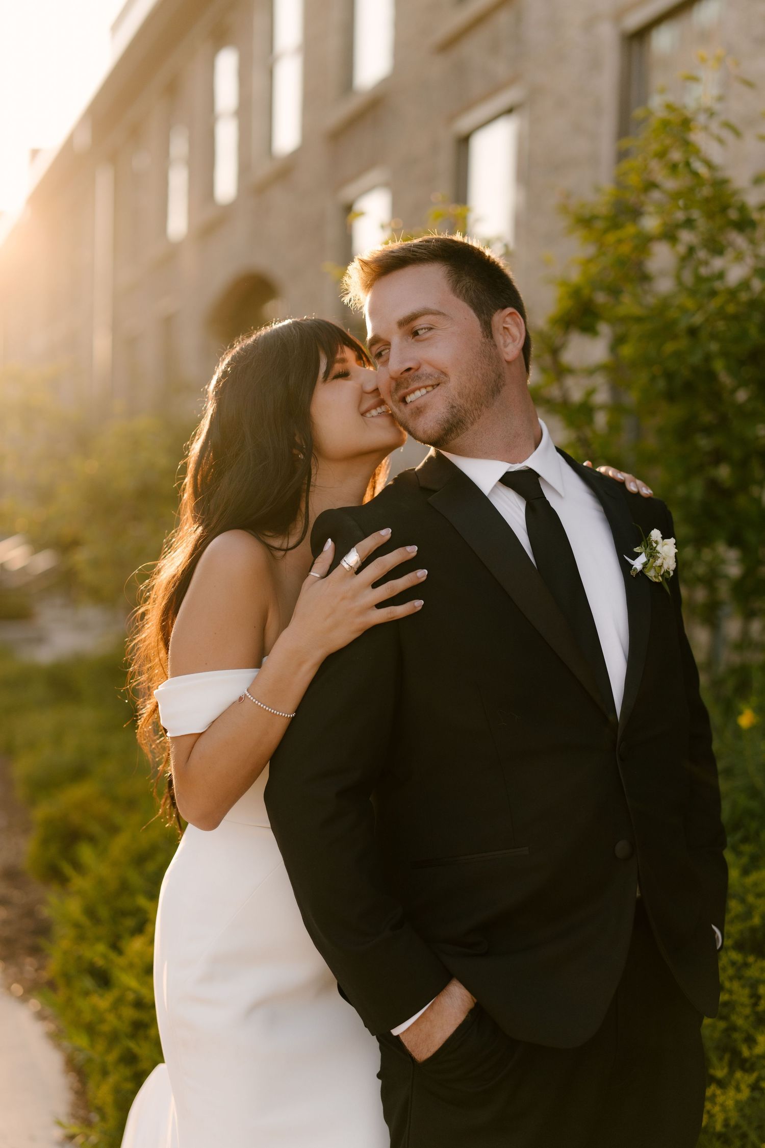 Bride kissing groom on cheek at golden hour, both smiling outdoors near Tapestry Hall building with warm sunlight.