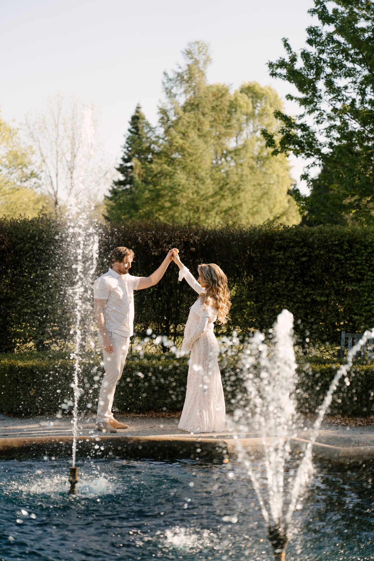 Couple in white dancing by a garden fountain with water jets sparkling in the sunlight.