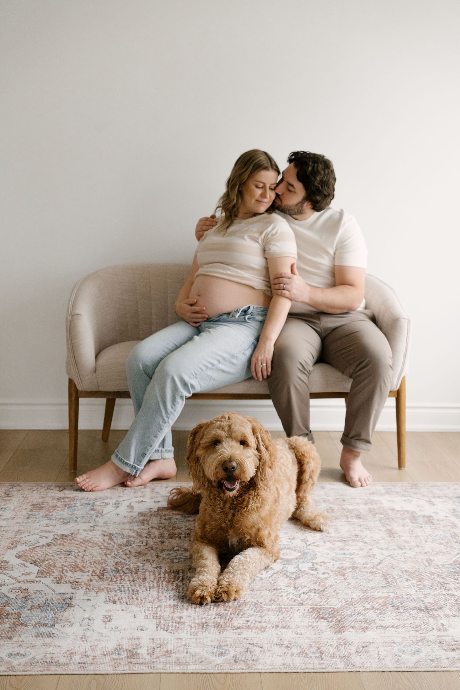A couple sits on a beige sofa with a golden doodle dog lying on the rug during a maternity session.