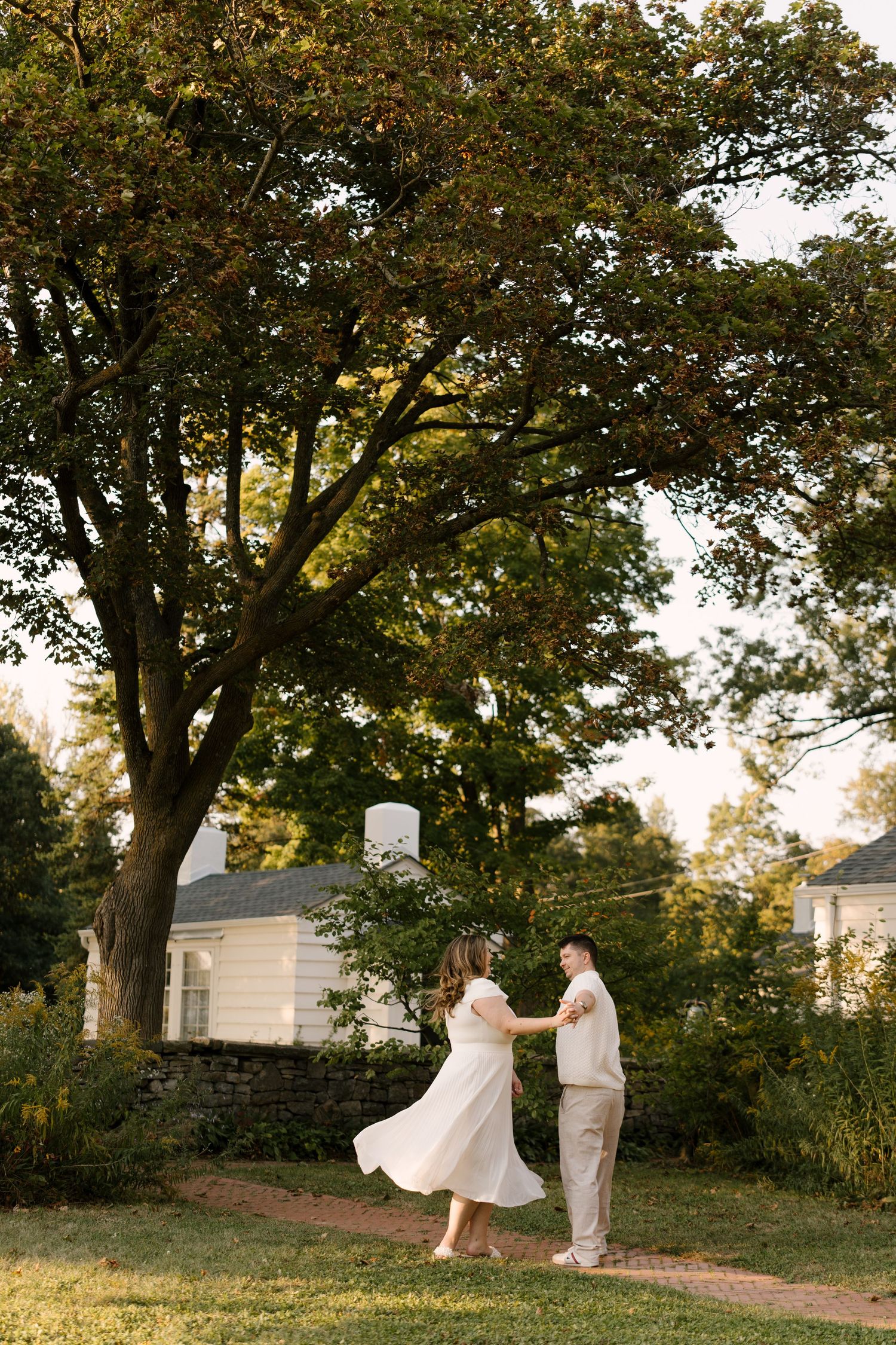 Couple dancing and twirling on a green lawn at golden hour with large trees and white buildings.