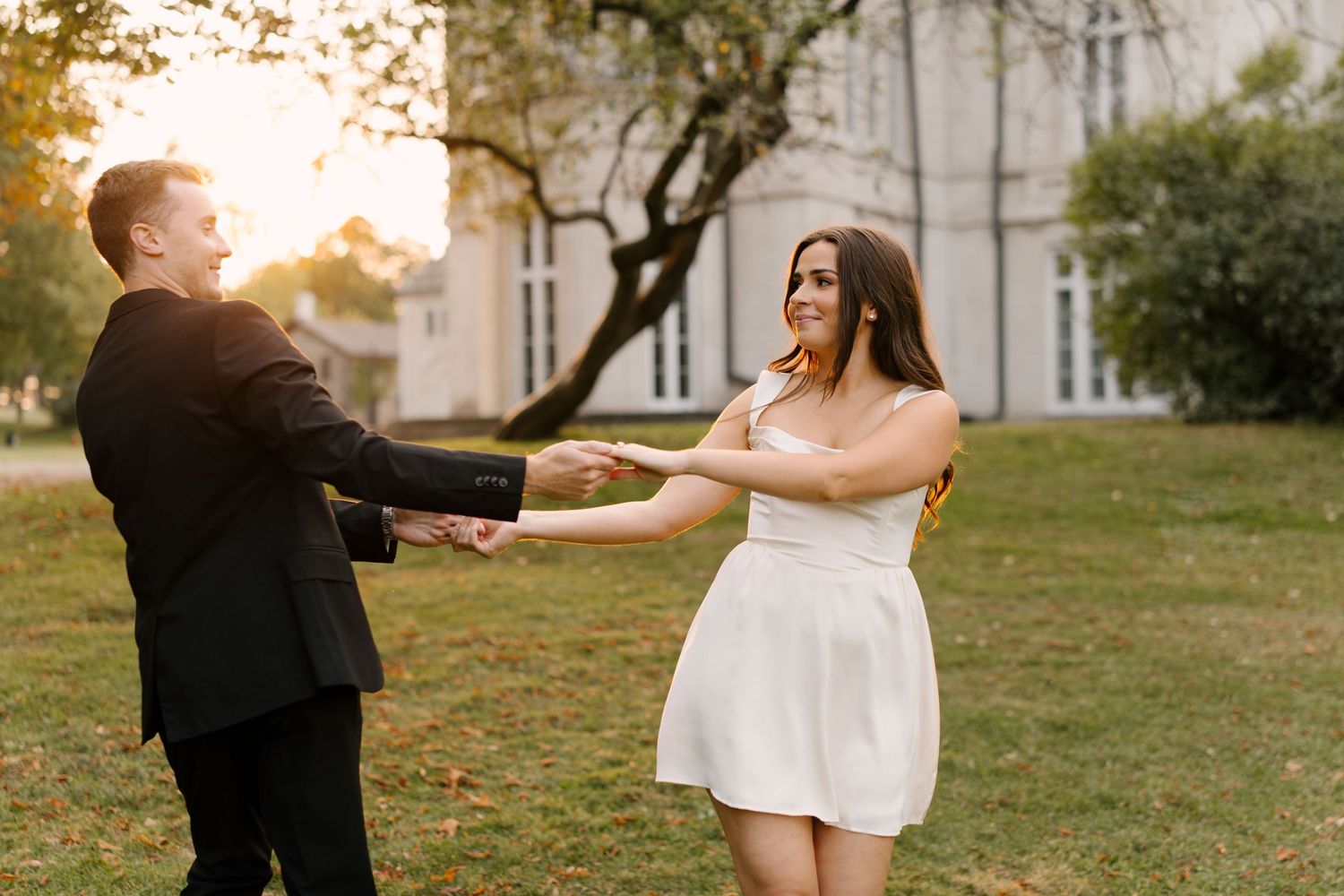 Couple dancing and holding hands on grass at golden hour with white manor house in background.