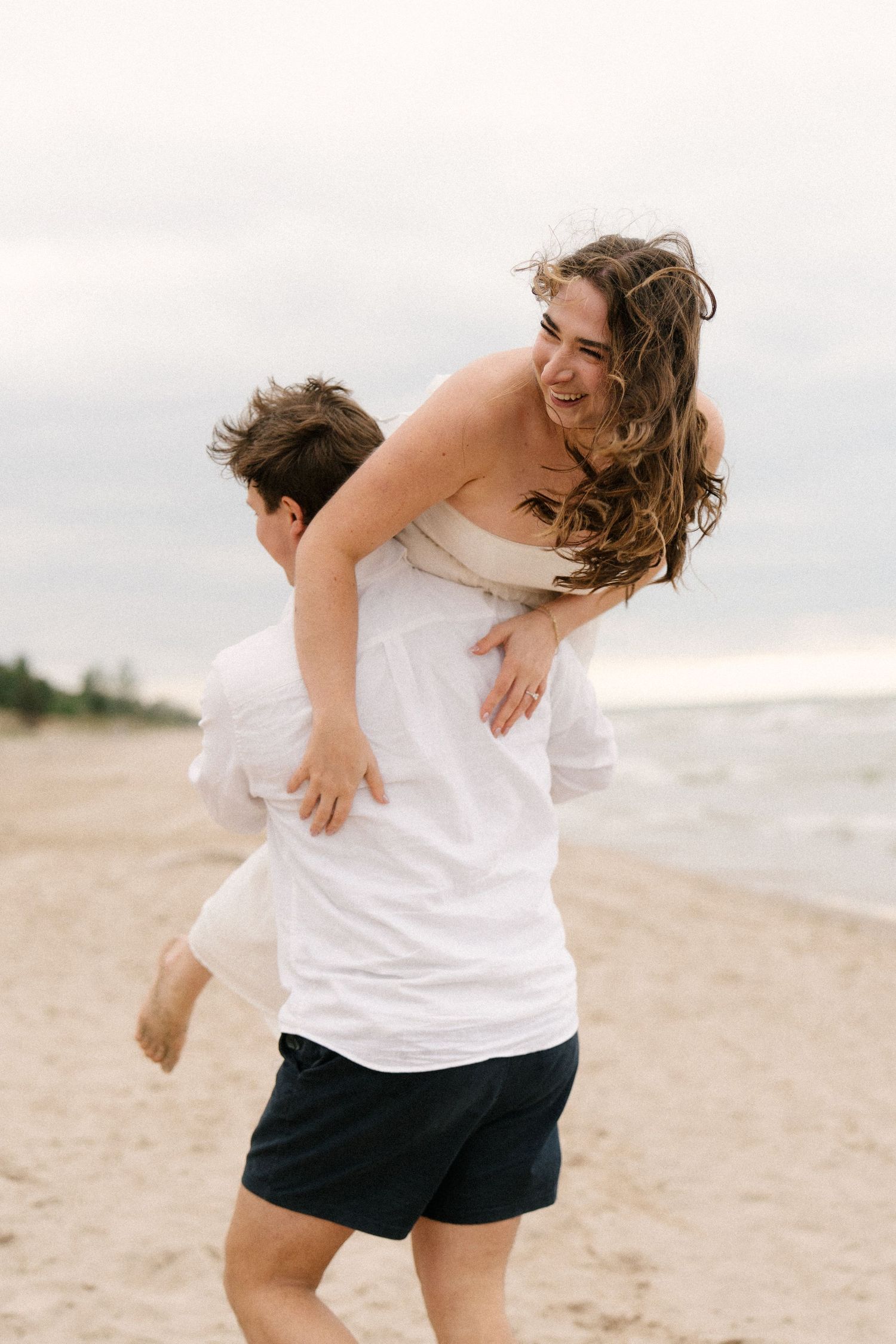Man giving laughing woman a piggyback ride on sandy beach, woman in white dress with hair blowing in wind.