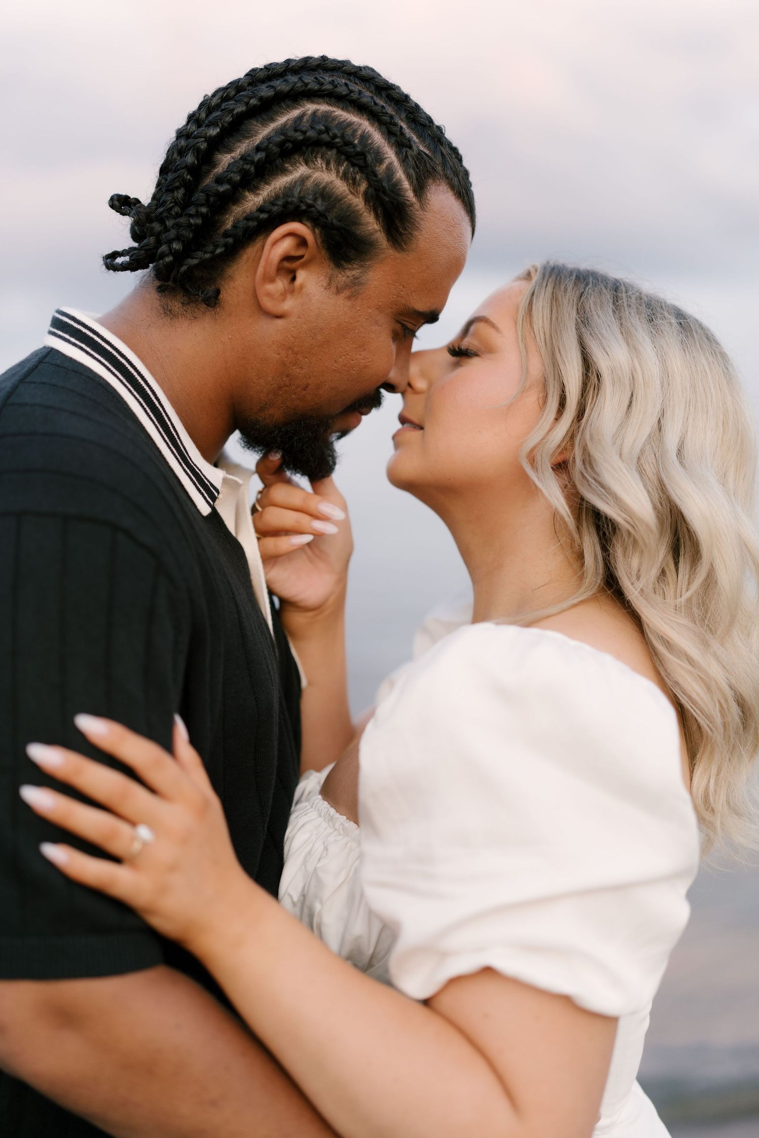 A couple shares an intimate almost-kiss moment outdoors, the woman wearing a white off-shoulder dress.
