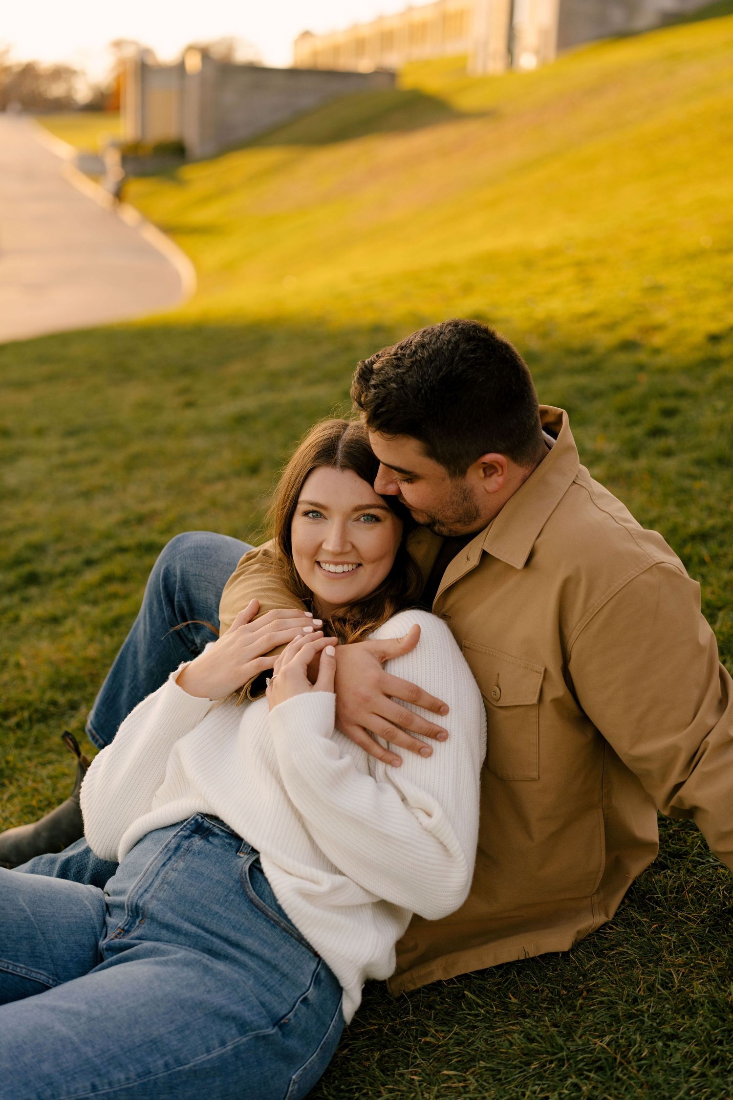 Happy couple cuddling on green grass at golden hour, man embracing smiling woman from behind outdoors.