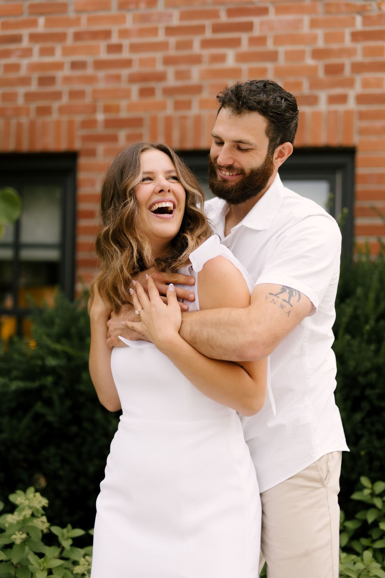 Bearded groom hugging laughing brunette fiancee in white dress in front of a brick building.