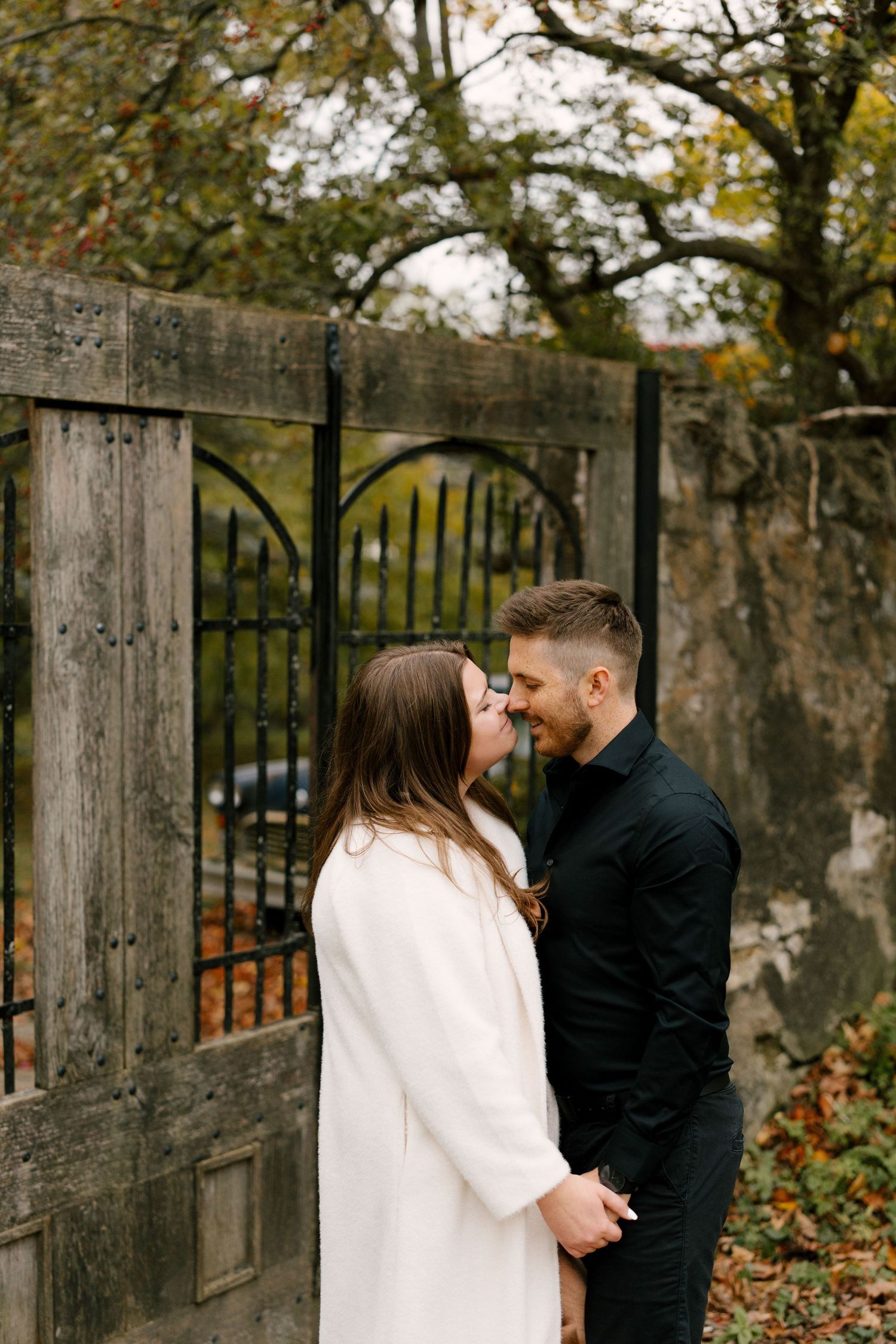 Couple sharing an intimate moment in front of a rustic wooden gate with autumn foliage in the background.