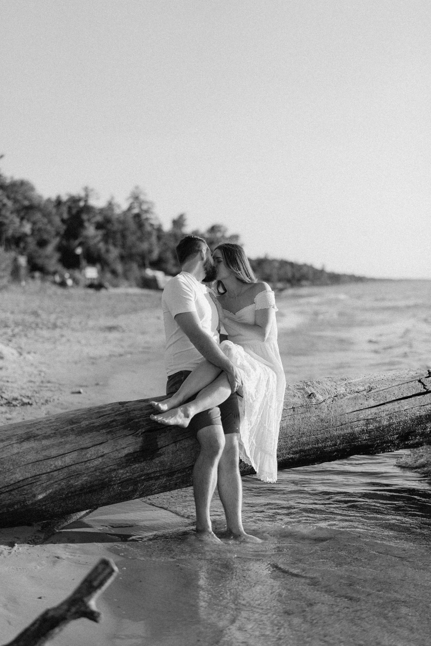 Black and white photo of couple kissing on a driftwood log at the water's edge.