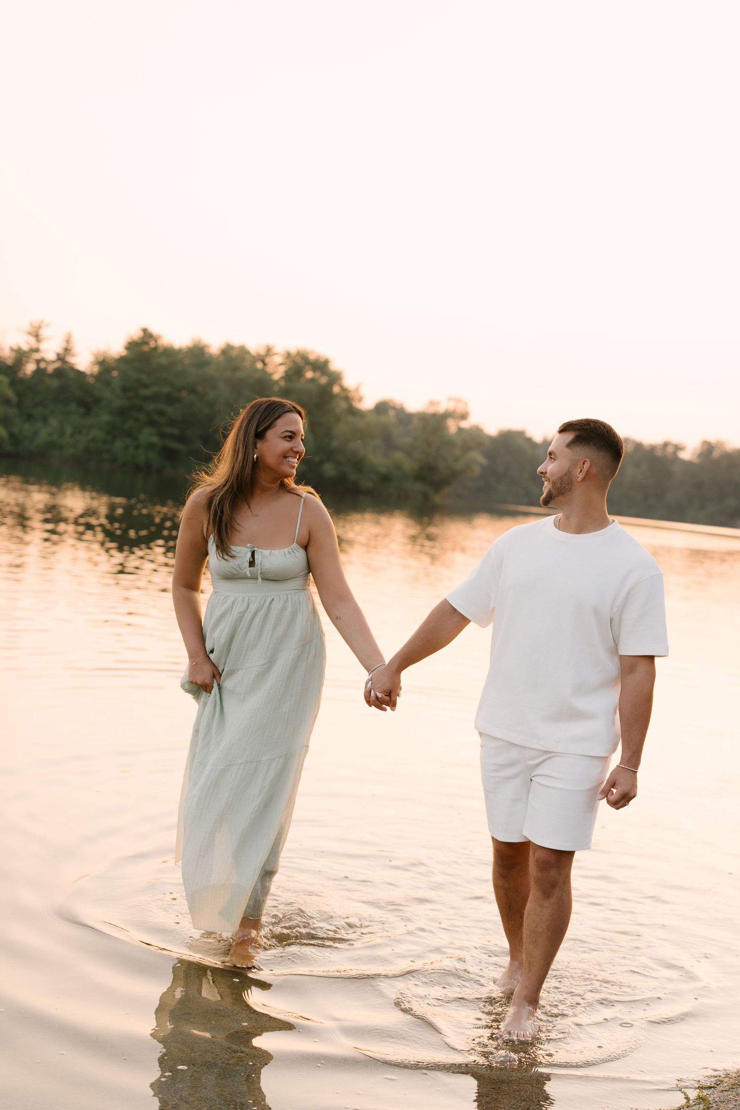 A smiling couple holds hands while wading through calm lake water together at a beautiful golden sunset.