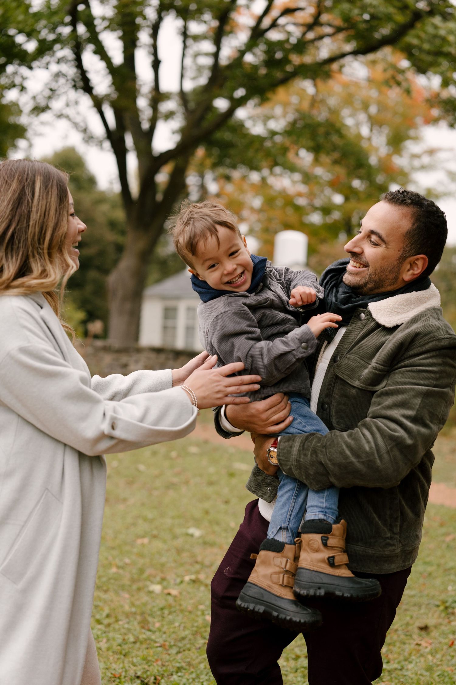 Happy family with pregnant mother, father holding toddler son, laughing outdoors in autumn park.