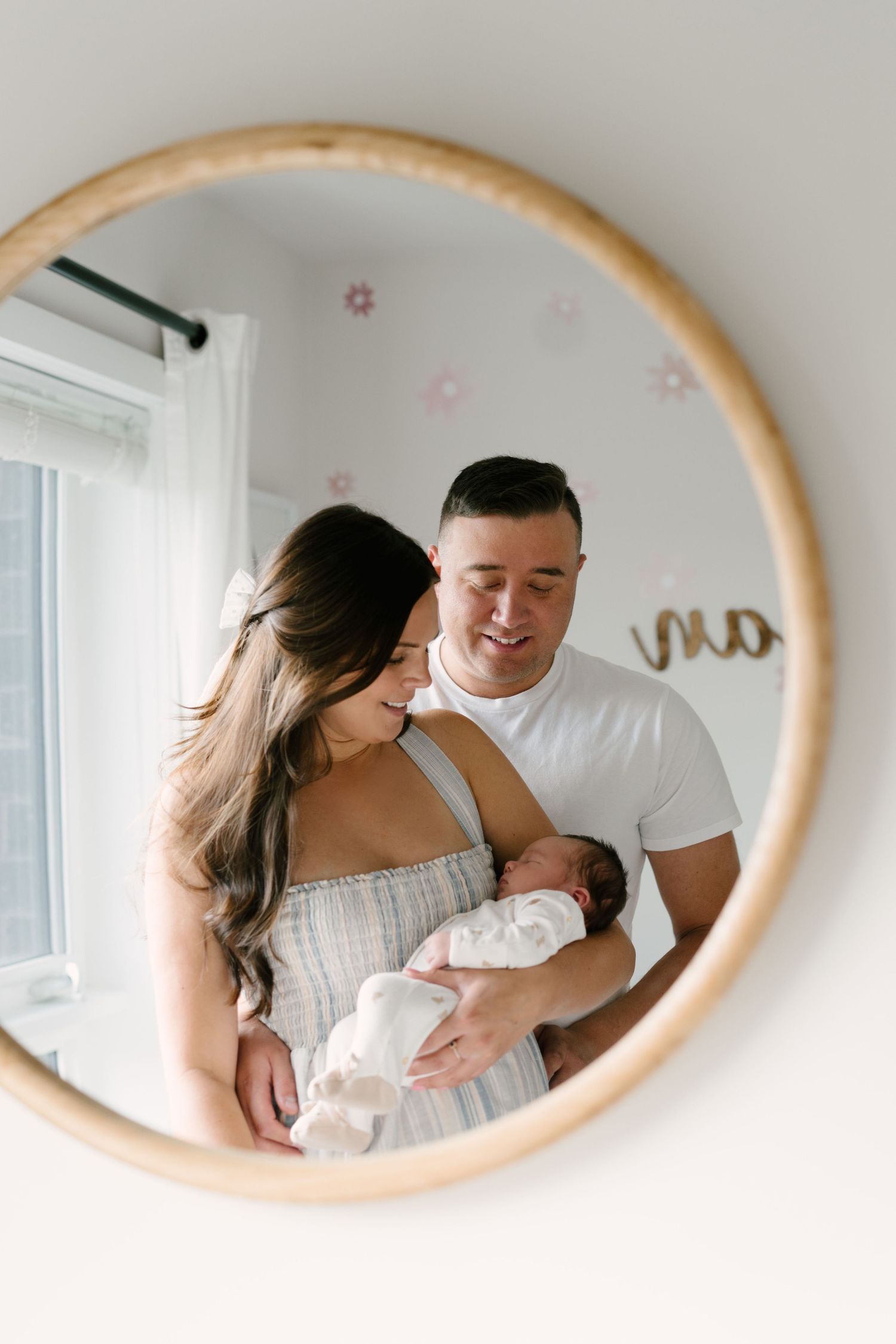 Happy couple with newborn baby girl reflected in round gold mirror in floral nursery.