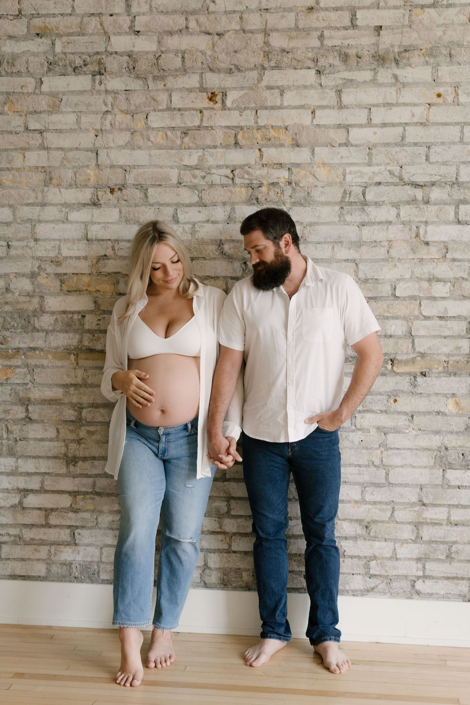 Pregnant couple holding hands against brick wall, woman in white bralette showing baby bump, man in white shirt and jeans.