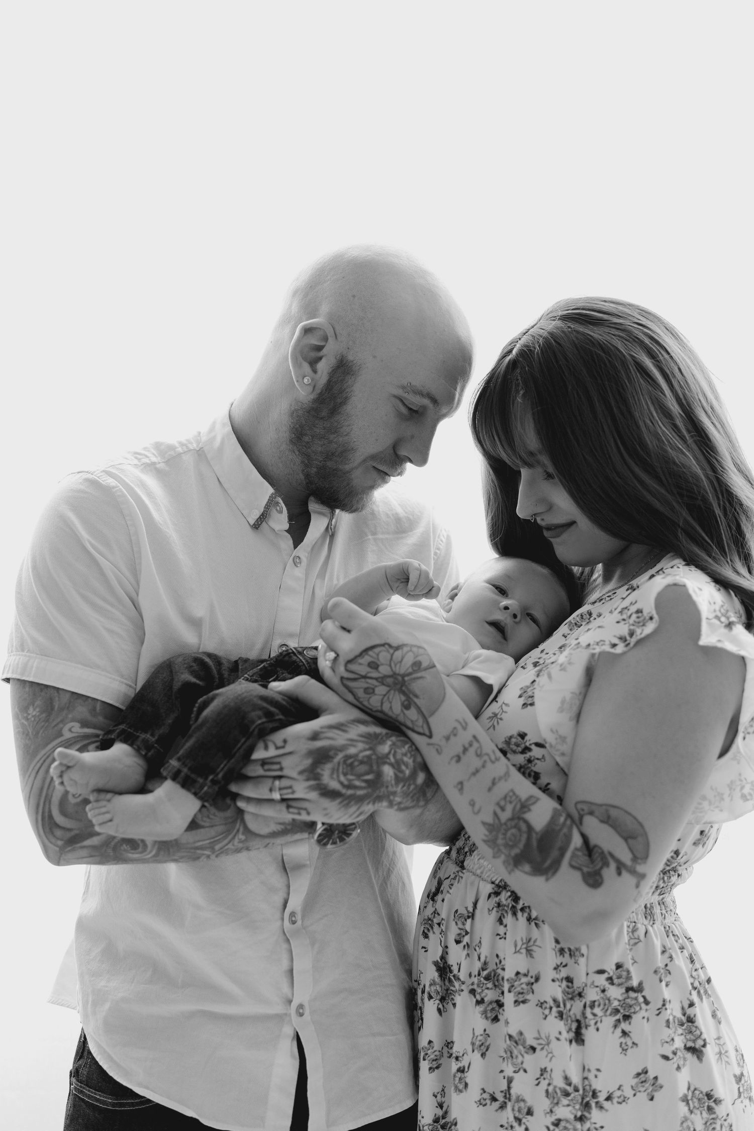 Black and white portrait of tattooed parents tenderly looking down at their sleeping newborn baby.