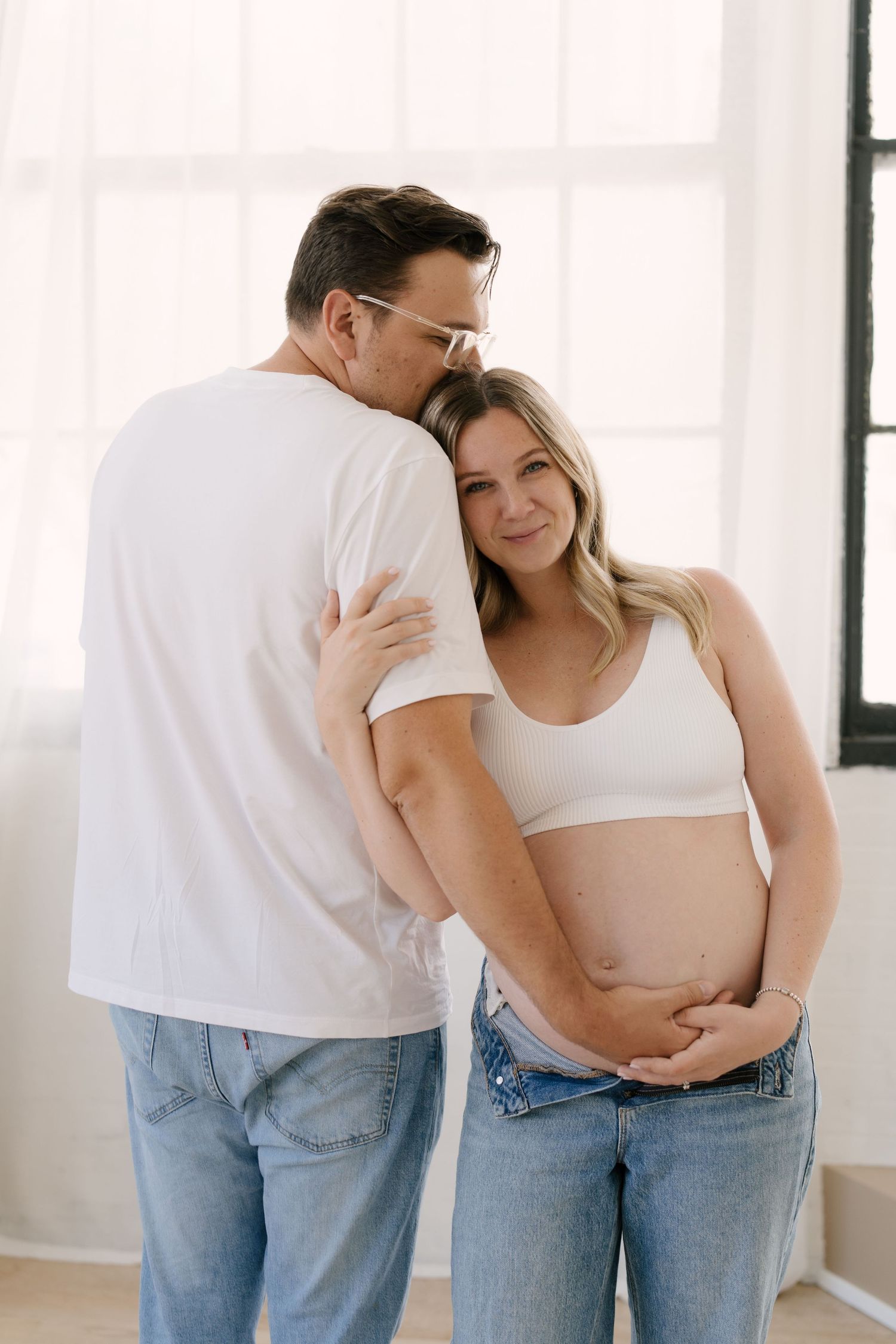 Couple embracing indoors, pregnant woman in white crop top and jeans smiling while partner kisses her head.